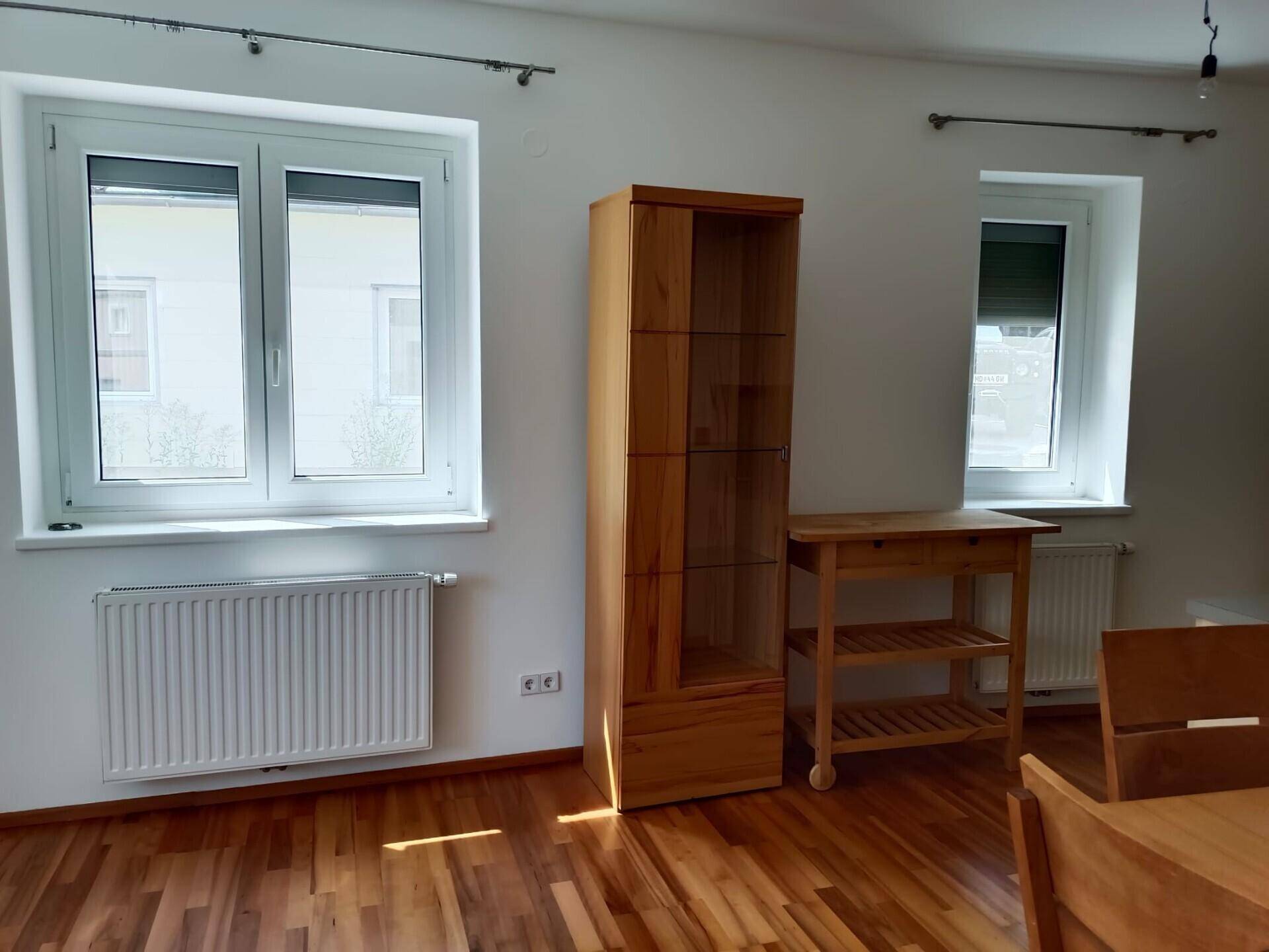 Wohn- und Essbereich mit Holzmöbeln, zwei Fenstern und viel Tageslicht.