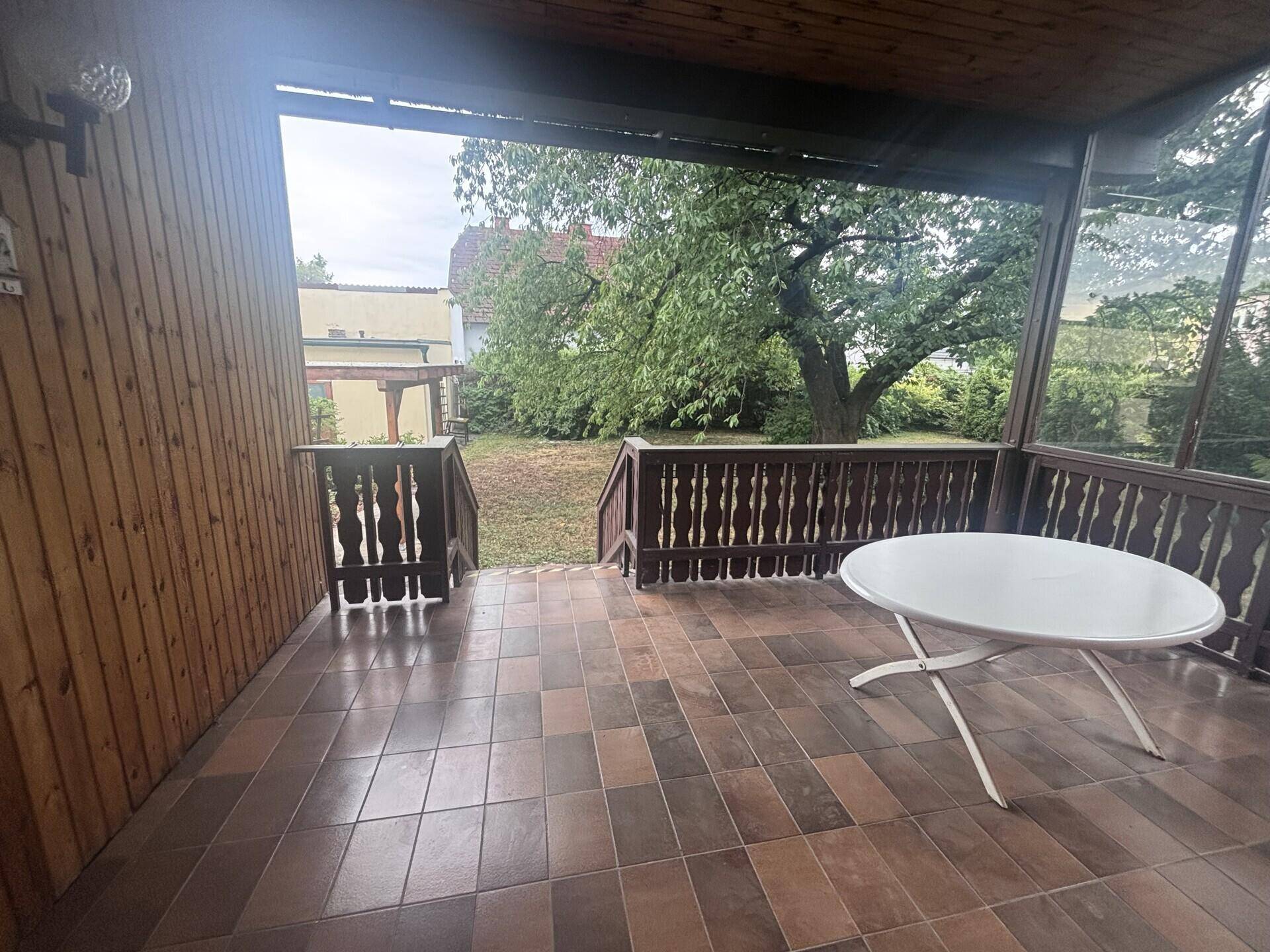 Überdachte Terrasse mit braunen Fliesen und einem Tisch, mit Blick auf den grünen Garten.