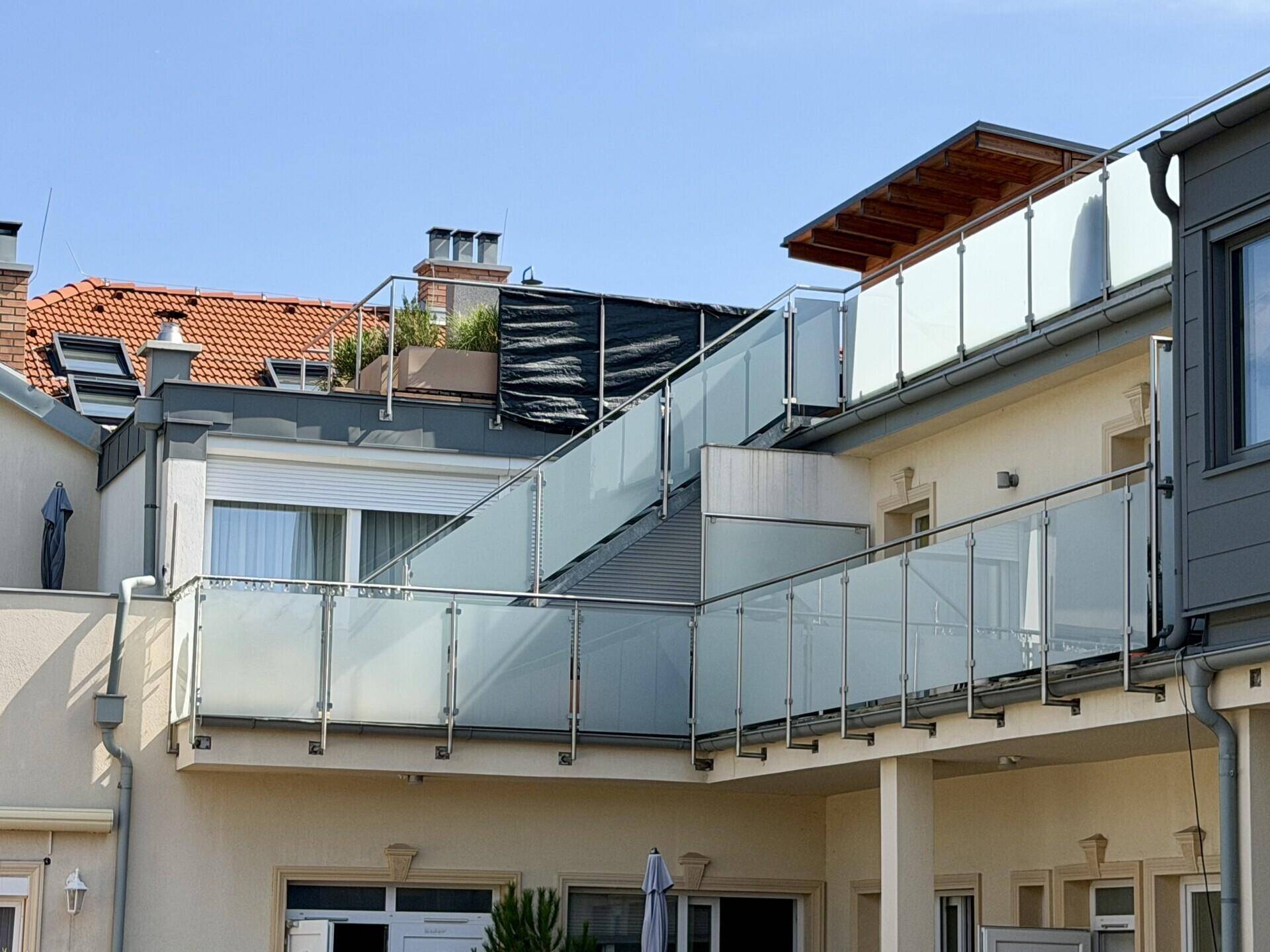 Mehrere Terrassen und Balkone mit modernen Glasgeländern an der Gebäudefassade unter blauem Himmel.