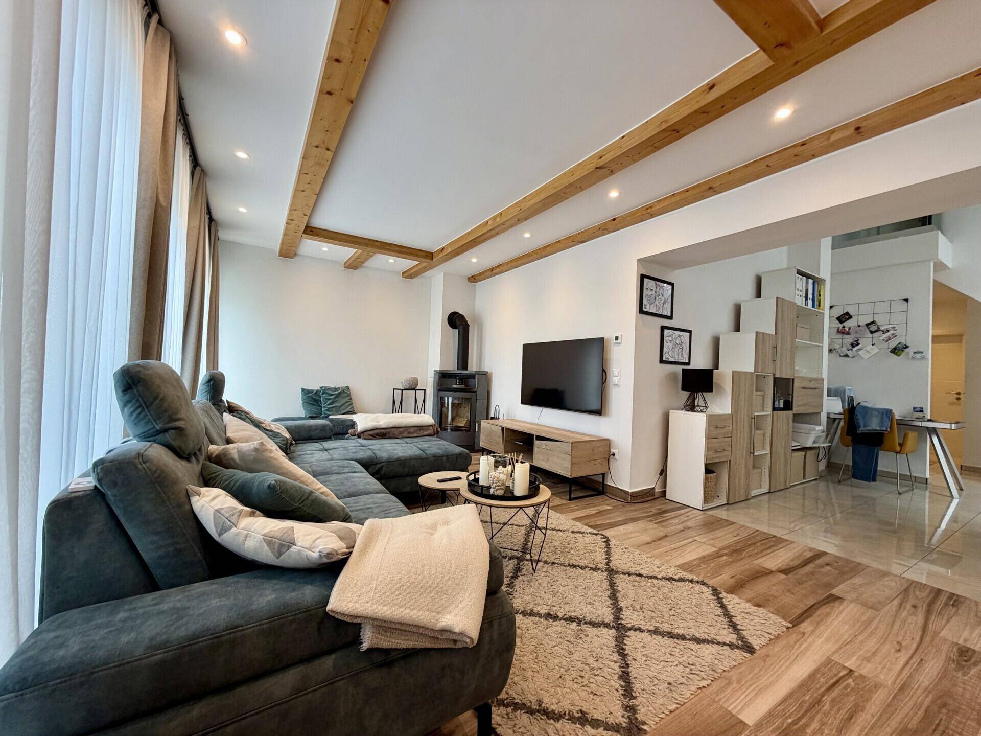 Modernes Wohnzimmer mit großem Sofa, Holzbalken und Kaminofen, offen zum Essbereich.