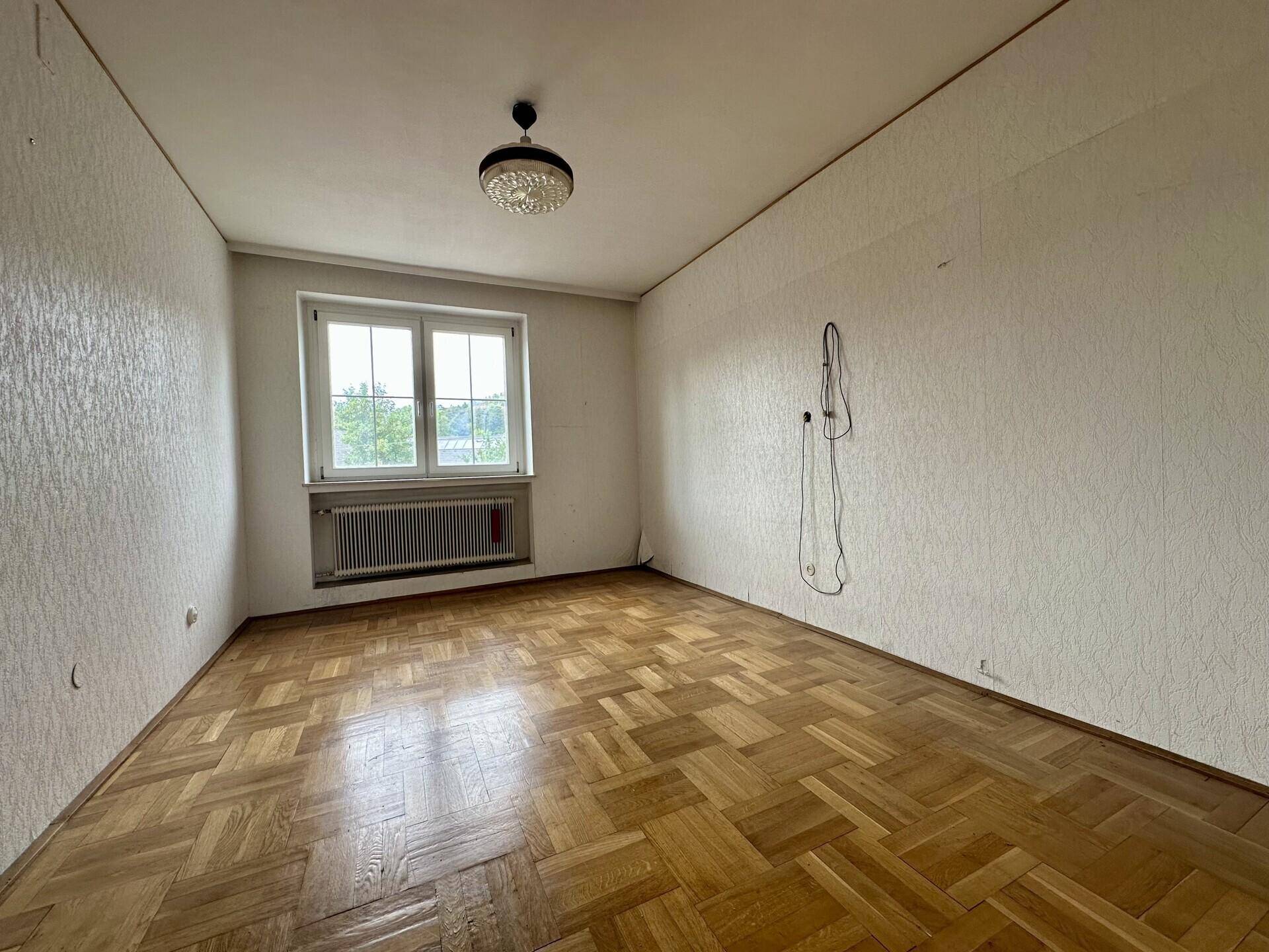 Schlafzimmer mit Parkettboden, weißer Tapete und Fenster mit Blick nach draußen.