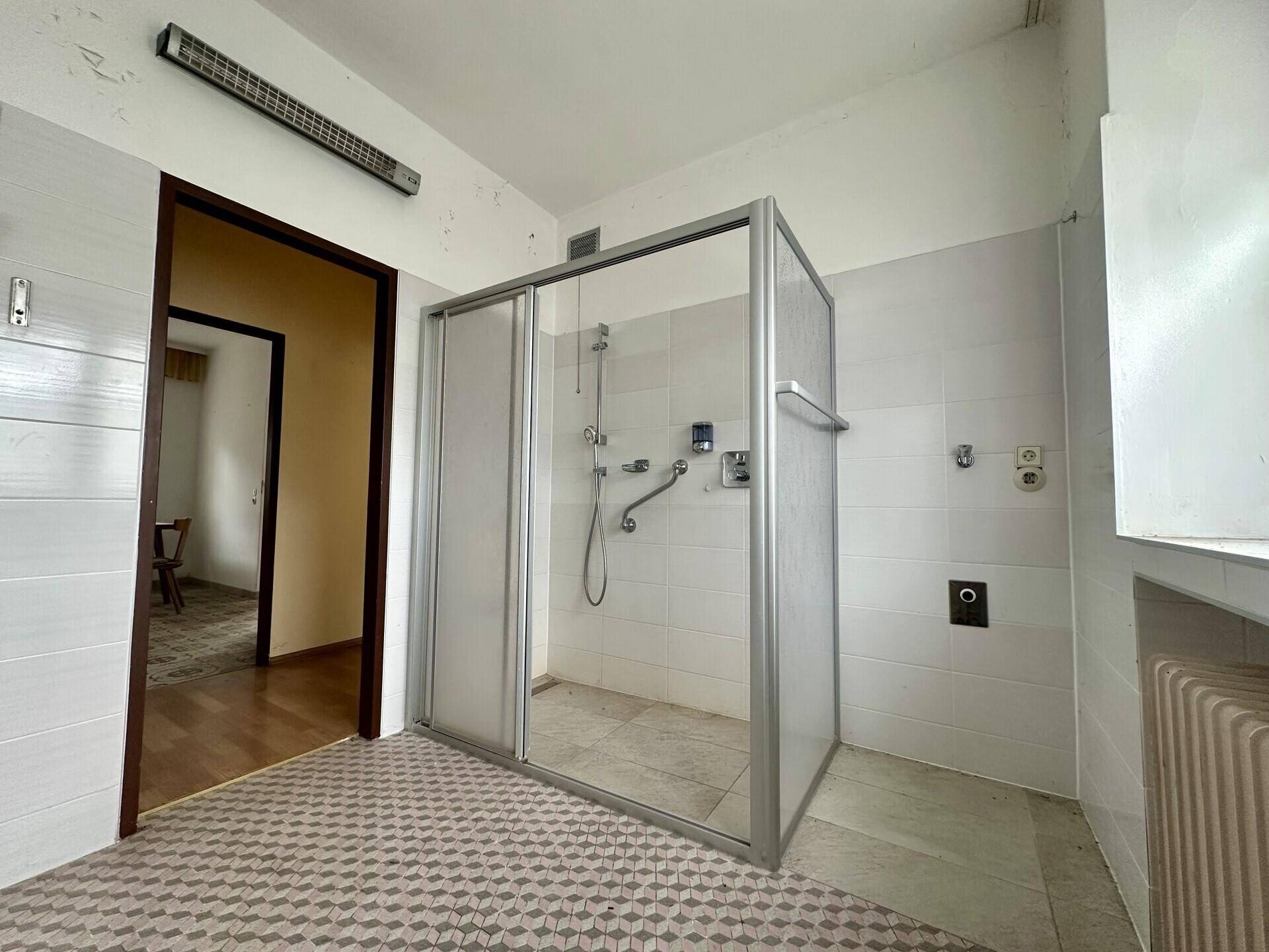 Badezimmer mit Duschkabine und gemusterten Fliesen, angrenzend an einen weiteren Raum.