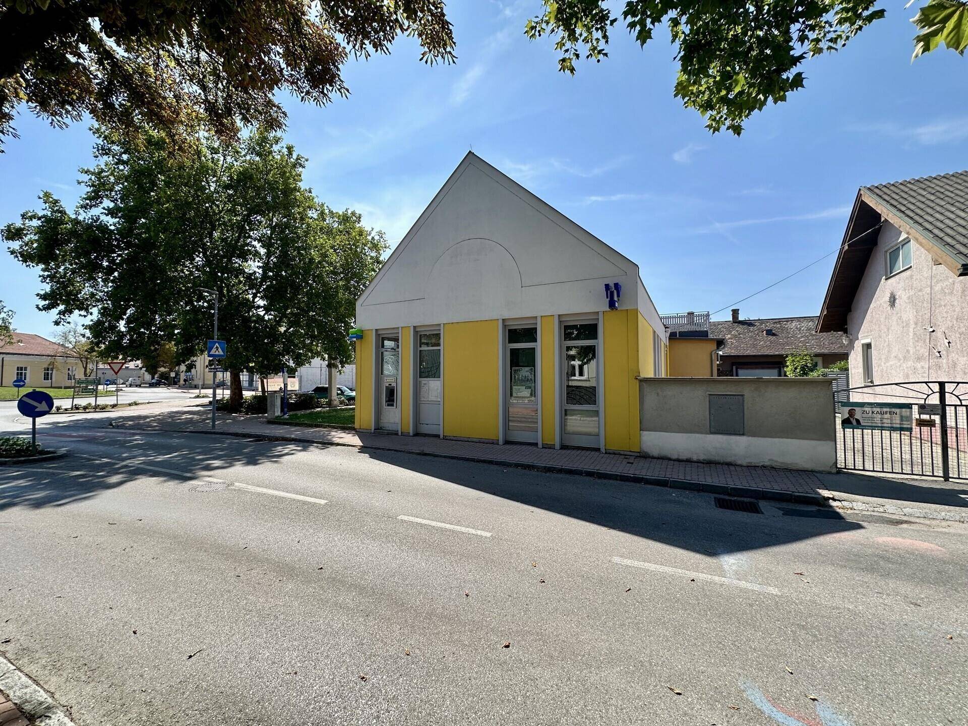 Seitenansicht des gelben Gebäudes an einer Straße mit weiteren Häusern und Bäumen im Hintergrund.