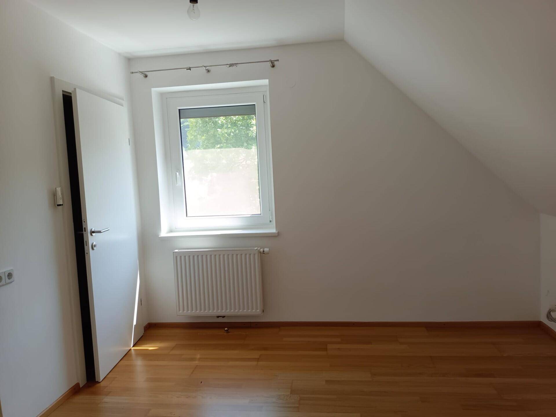 Helles Schlafzimmer im Dachgeschoss mit schrägen Wänden, Holzparkett und einem Fenster.