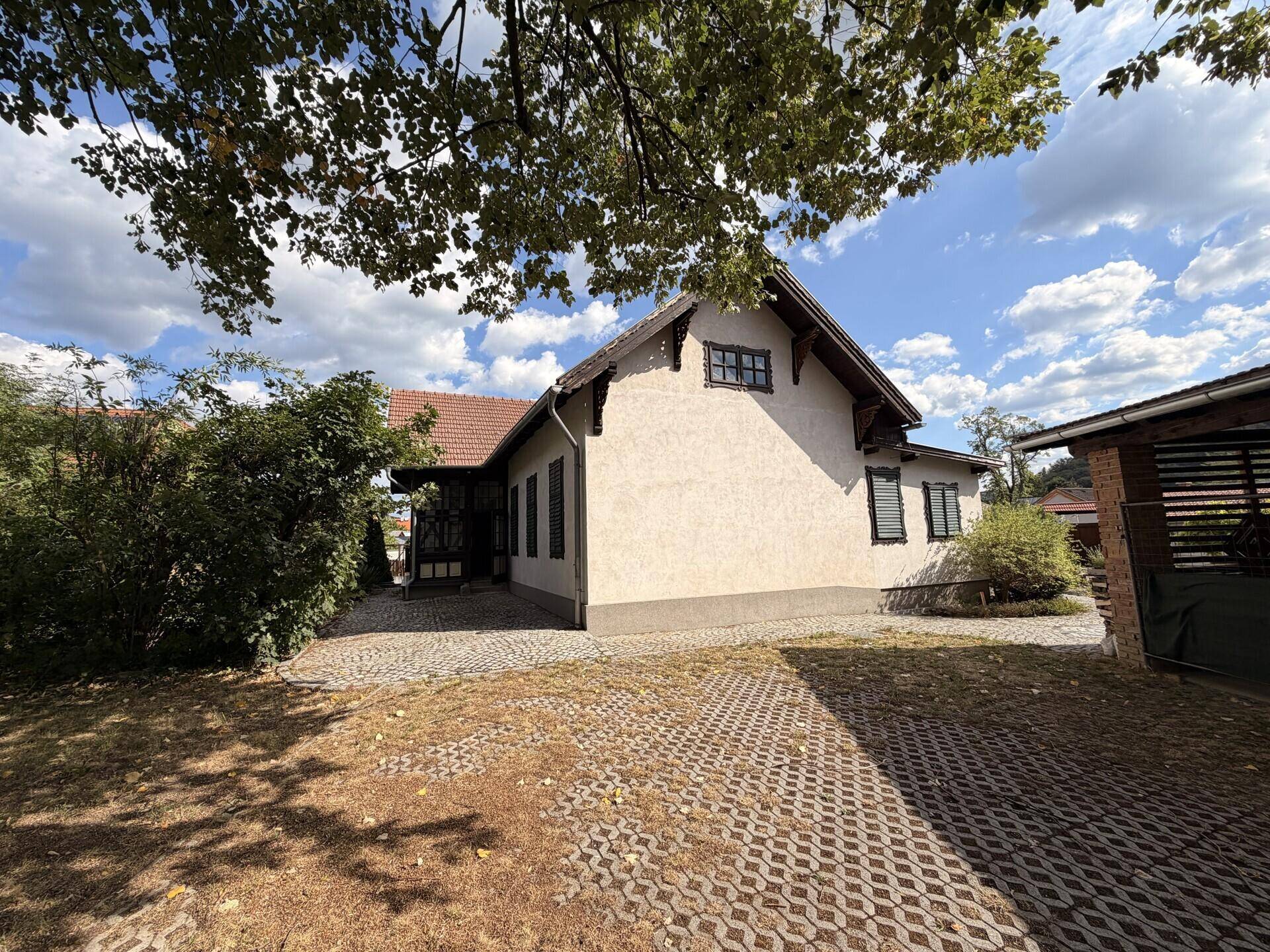 Seitenansicht eines traditionellen Hauses mit gepflasterter Auffahrt und altem Baumbestand im Vordergrund.