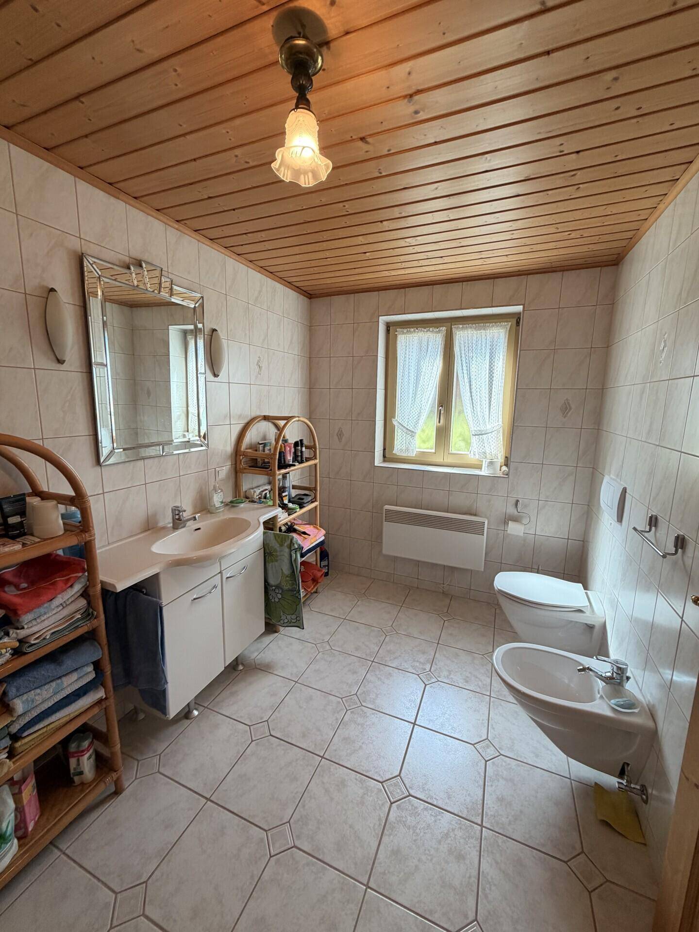 Helles Badezimmer mit Fliesenboden, Holzdecke, Waschbecken, Toilette und Bidet, sowie einem Fenster.