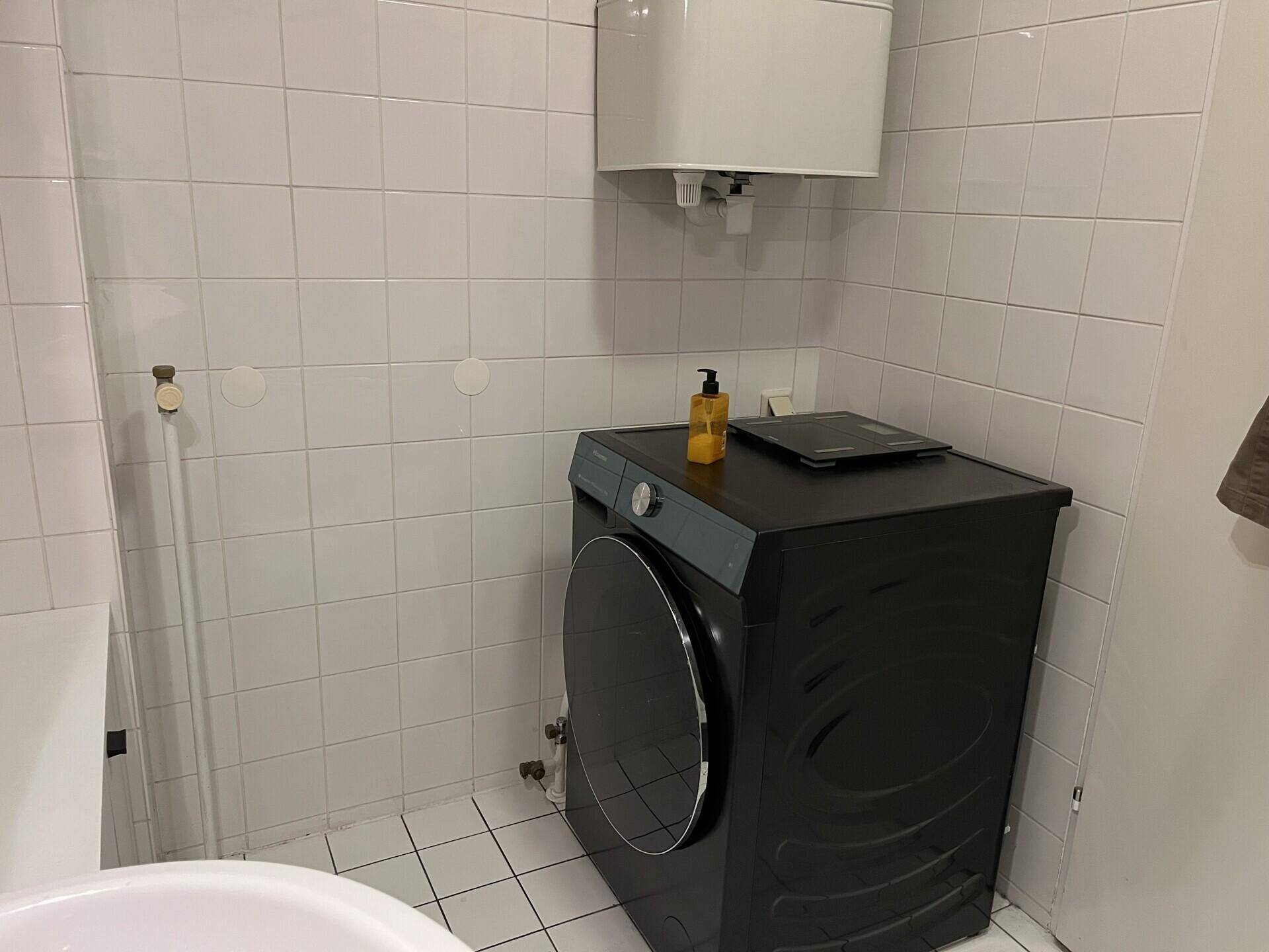 Badezimmer mit Waschmaschine und Warmwasserboiler, vollständig mit weißen Fliesen ausgestattet.