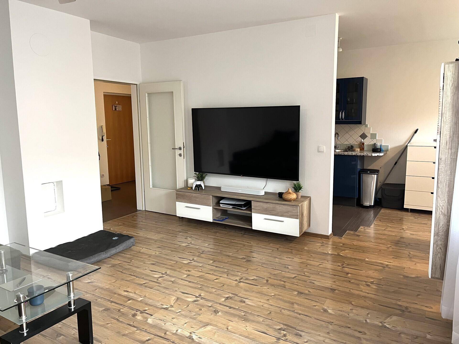 Wohnzimmer mit großem Wandfernseher und offenem Übergang zur Küche, mit Holzboden.