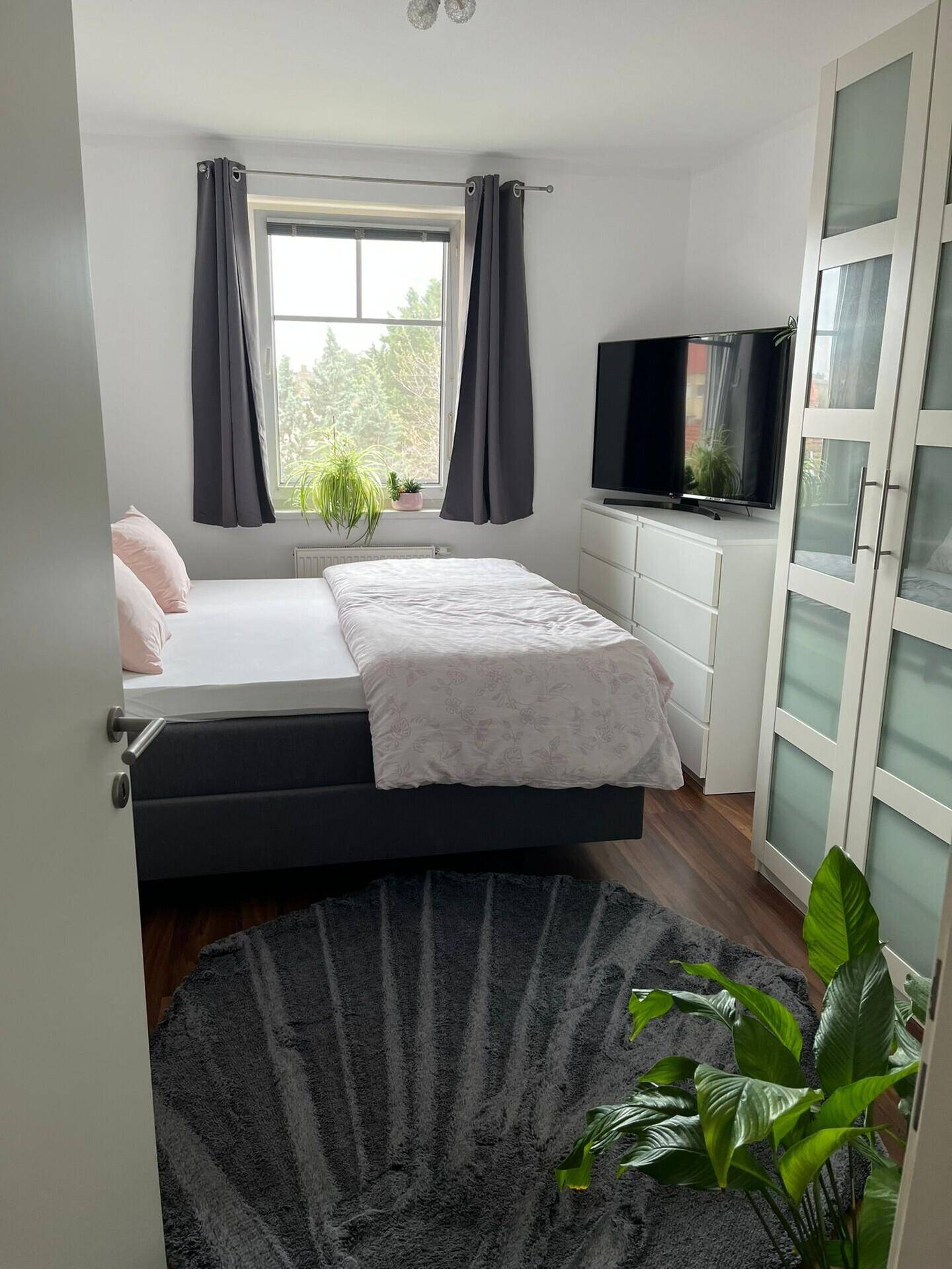 Helles Schlafzimmer mit Doppelbett, Kleiderschrank und Fernseher, mit Blick ins Grüne.
