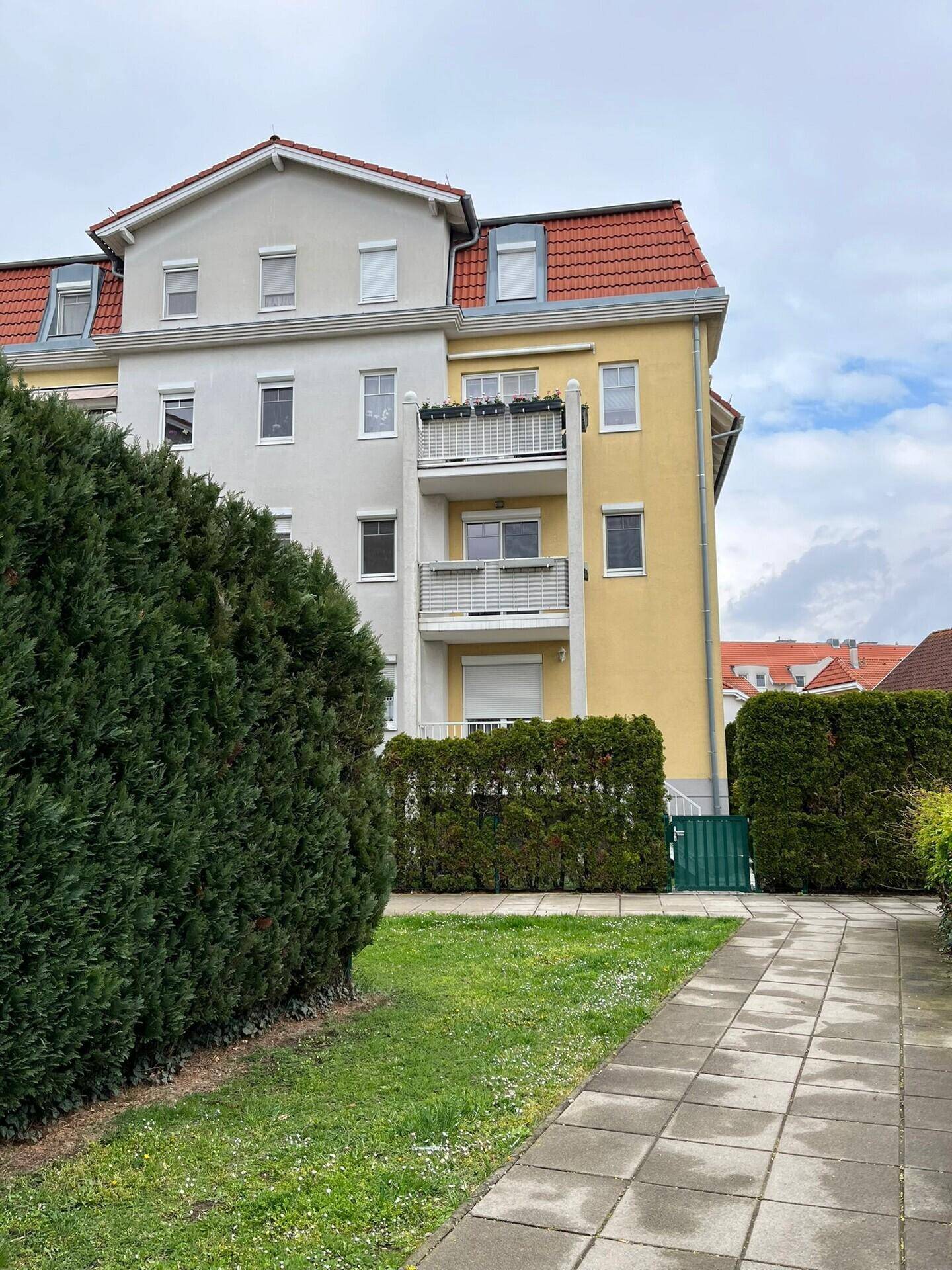 Mehrfamilienhaus mit gelber und grauer Fassade, roten Dächern und Balkonen, umgeben von Hecken.