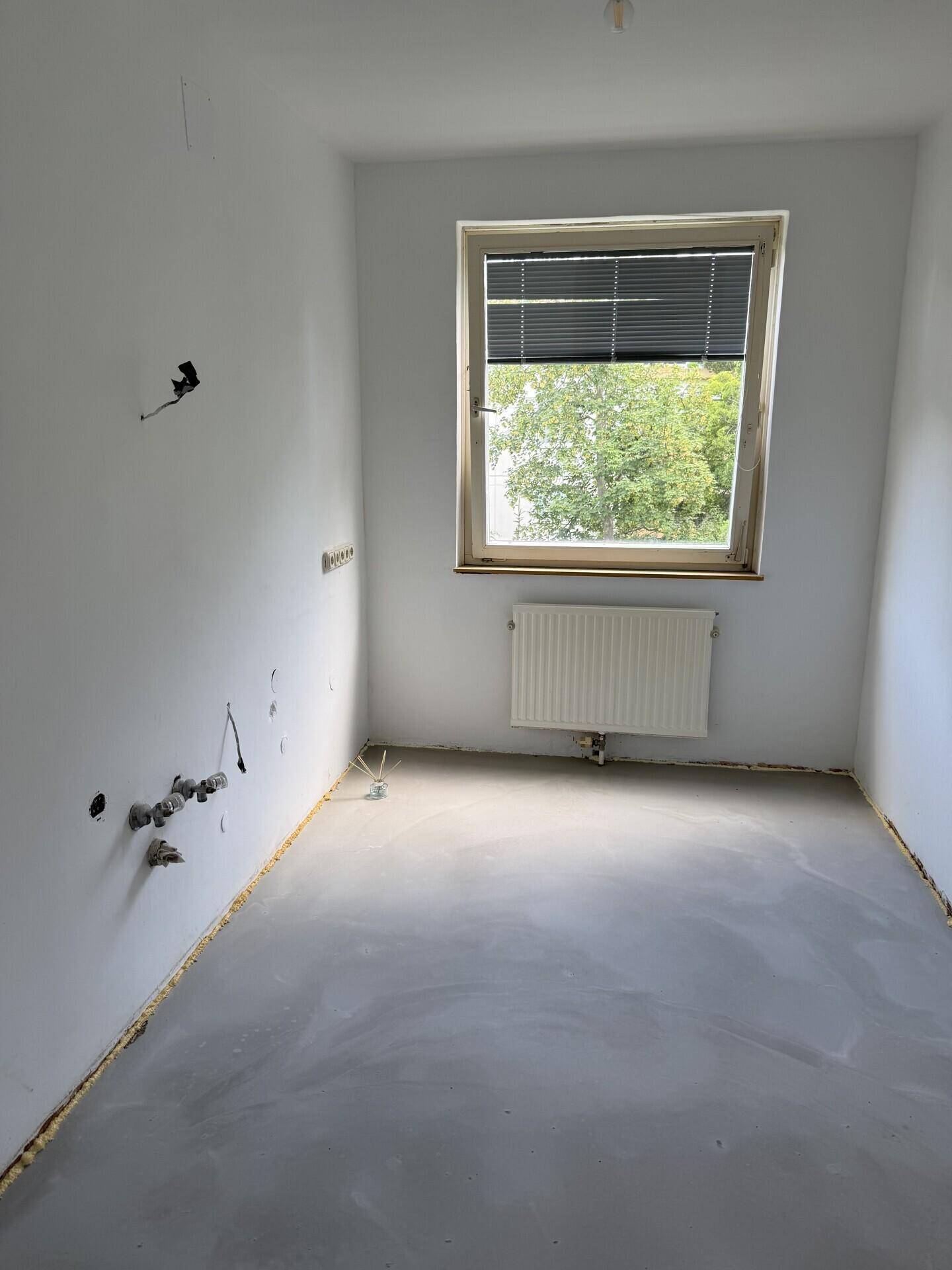 Leerer Raum mit Fenster, Heizkörper und sichtbaren Wasseranschlüssen für eine zukünftige Küche.