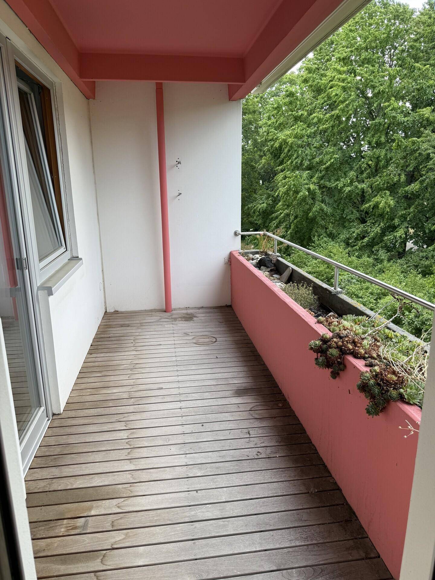 Langer Balkon mit Holzboden und rosa Brüstung, zugänglich von einem Innenraum.