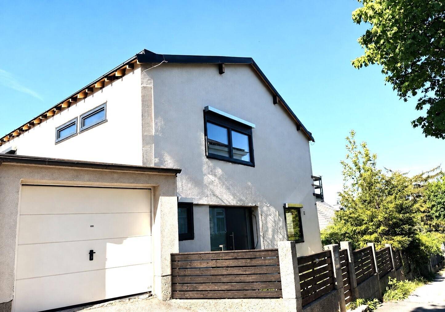Modernes beiges Einfamilienhaus mit Garage und Holzzaun unter blauem Himmel.