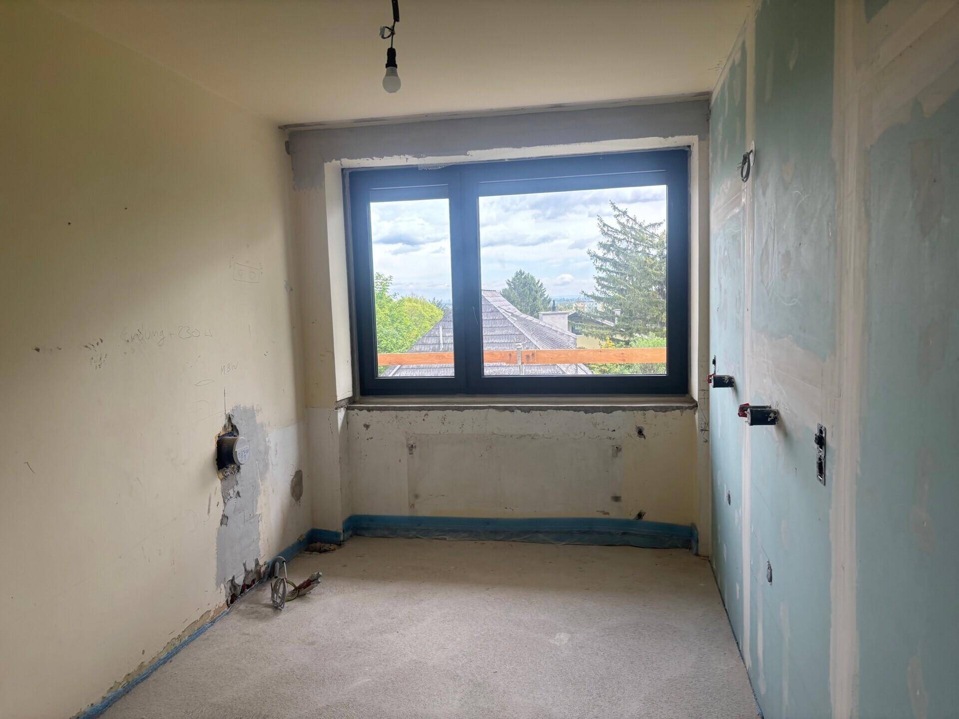 Unfertiger Raum im Rohbau mit einem Fenster und sichtbaren Installationen.