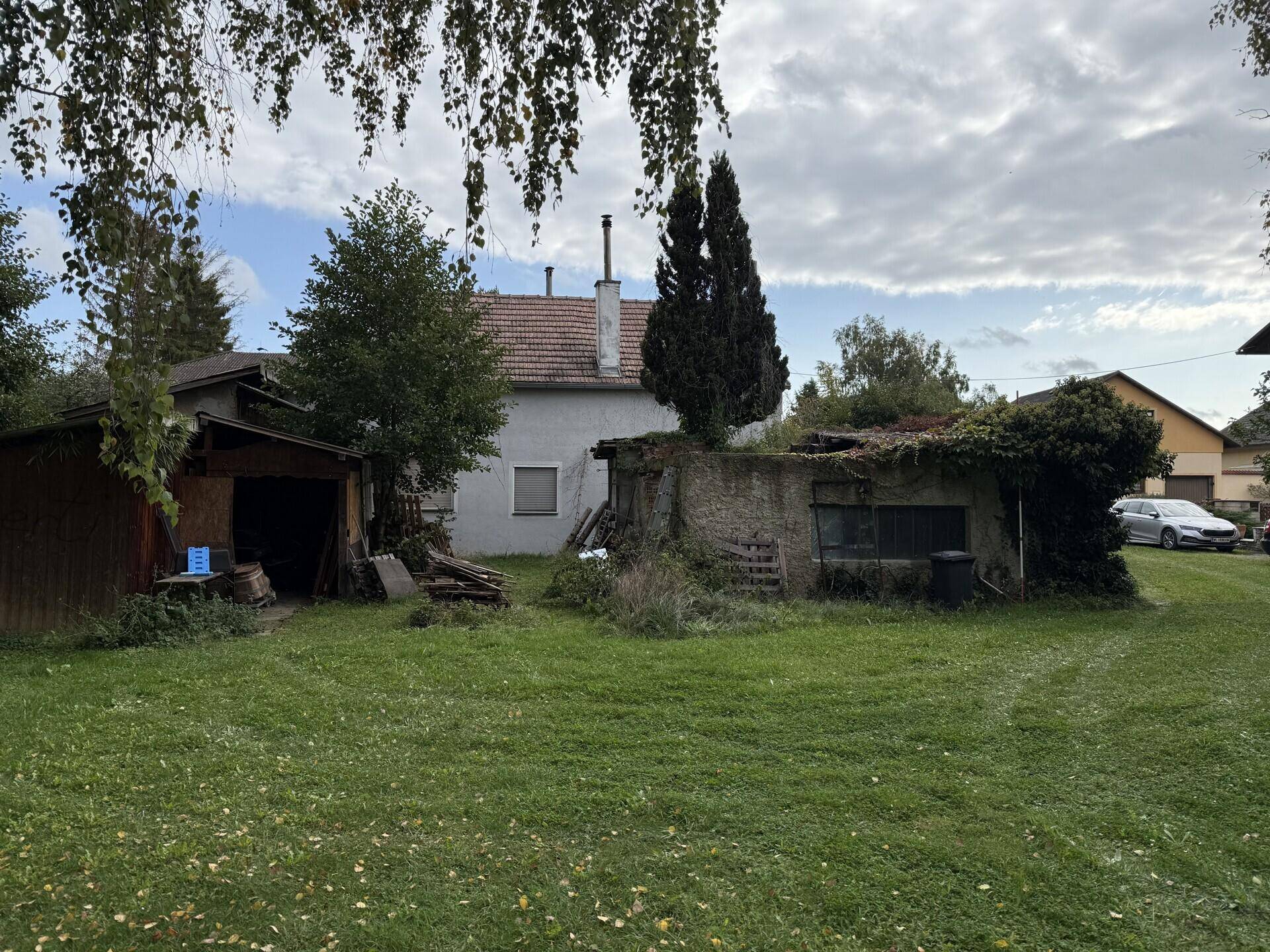 Hinterhofansicht mit altem Haus, Holzschuppen und einer überwucherten Ruine im Vordergrund.