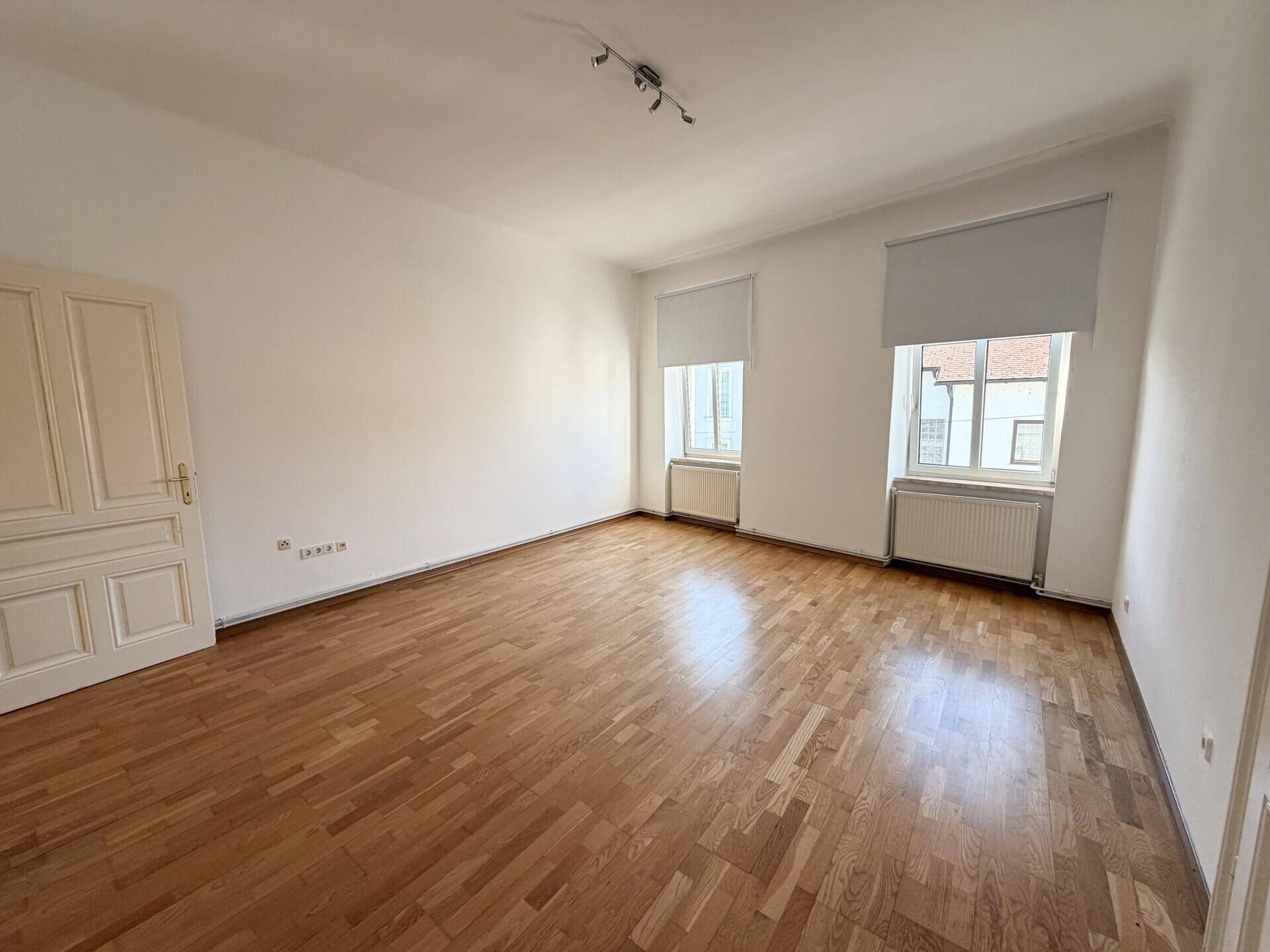 Geräumiges, helles Zimmer mit klassischem Parkettboden und zwei Fenstern, ideal für Wohnzwecke.