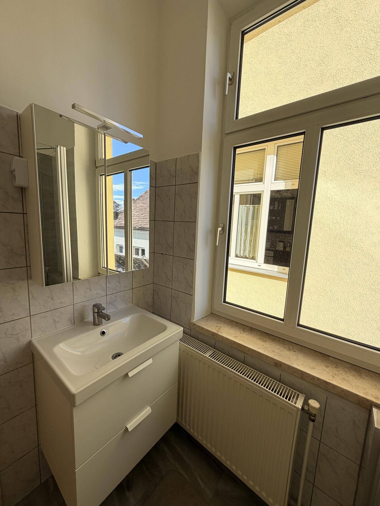 Helles Badezimmer mit Waschtisch, Spiegelschrank und großem Fenster, bietet viel Tageslicht.