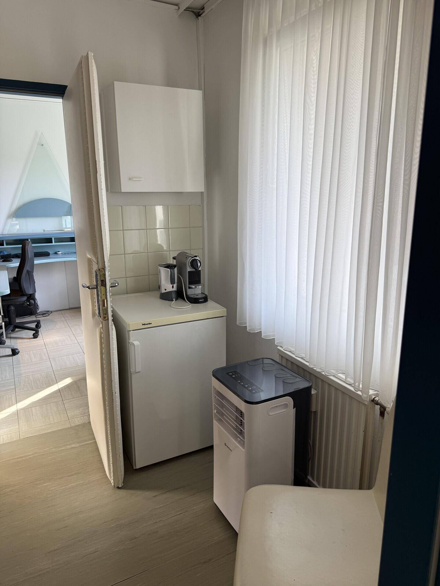 Kleine Küchenzeile mit Kühlschrank, Kaffeemaschine und einem Fenster mit weißen Vorhängen.