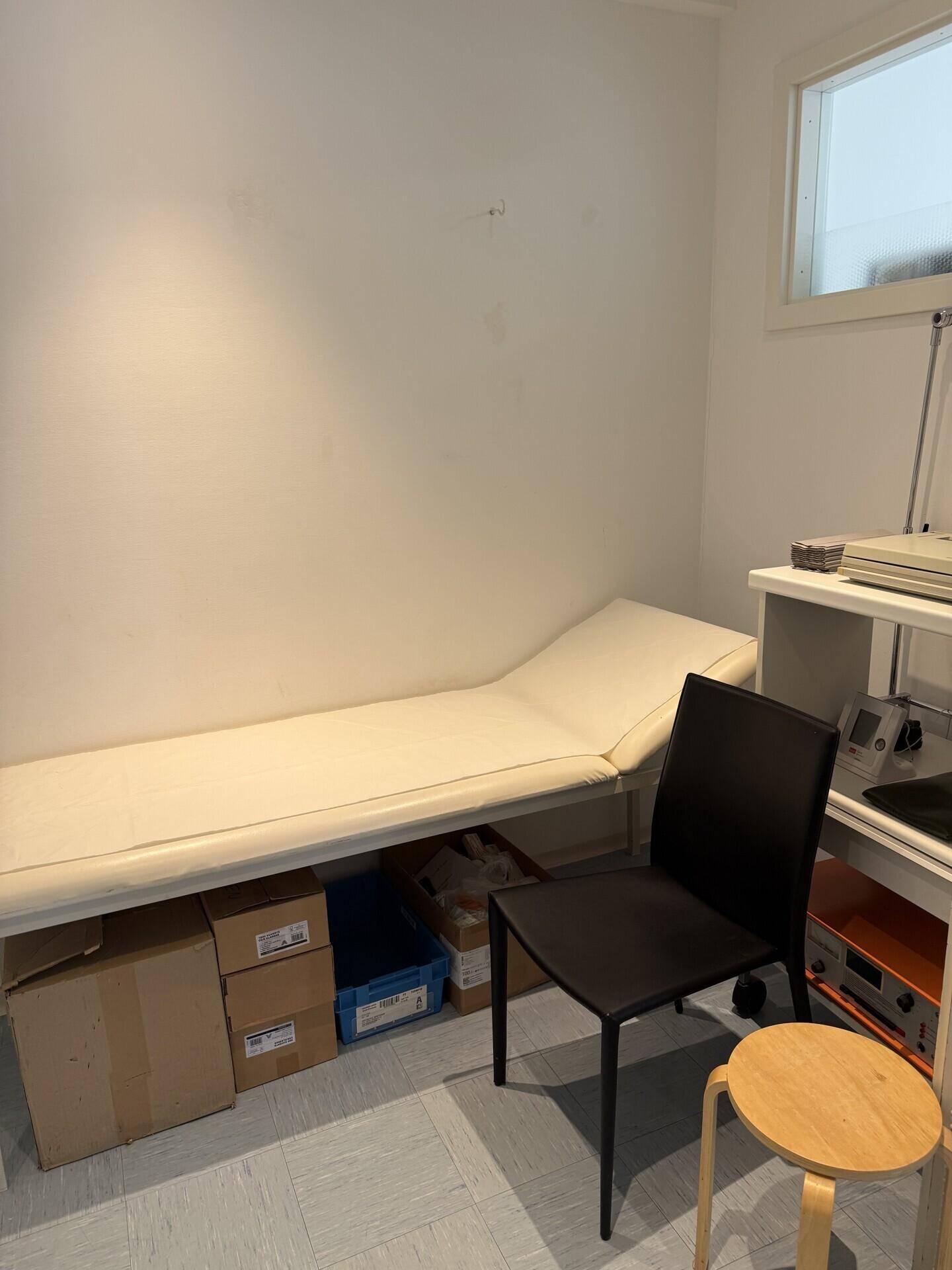 Kleines Behandlungszimmer mit Liege, schwarzem Stuhl und einem Fenster für natürliches Licht.