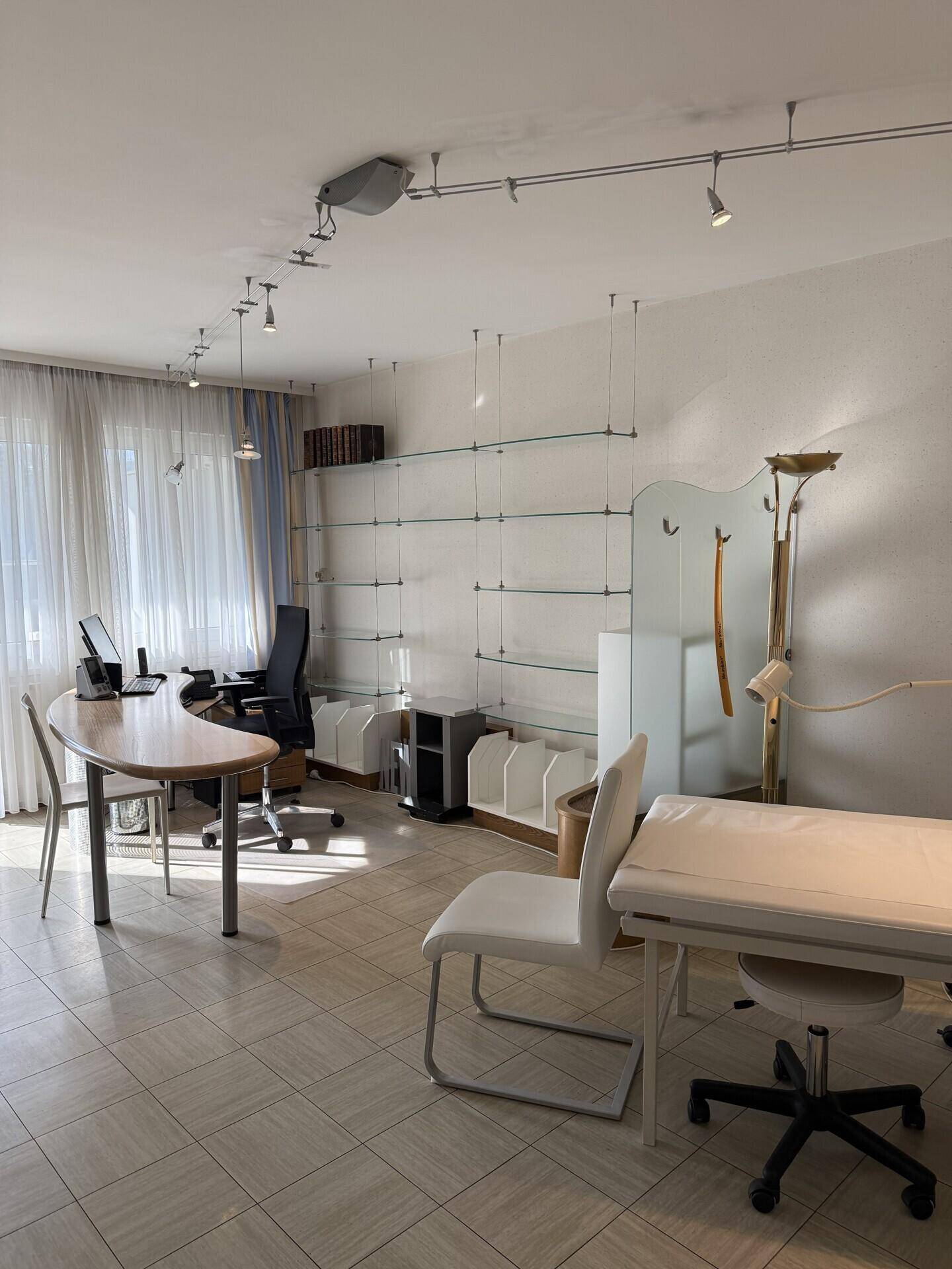 Behandlungszimmer oder Büro mit Untersuchungsliege, Schreibtisch und Glasregalen an der Wand.