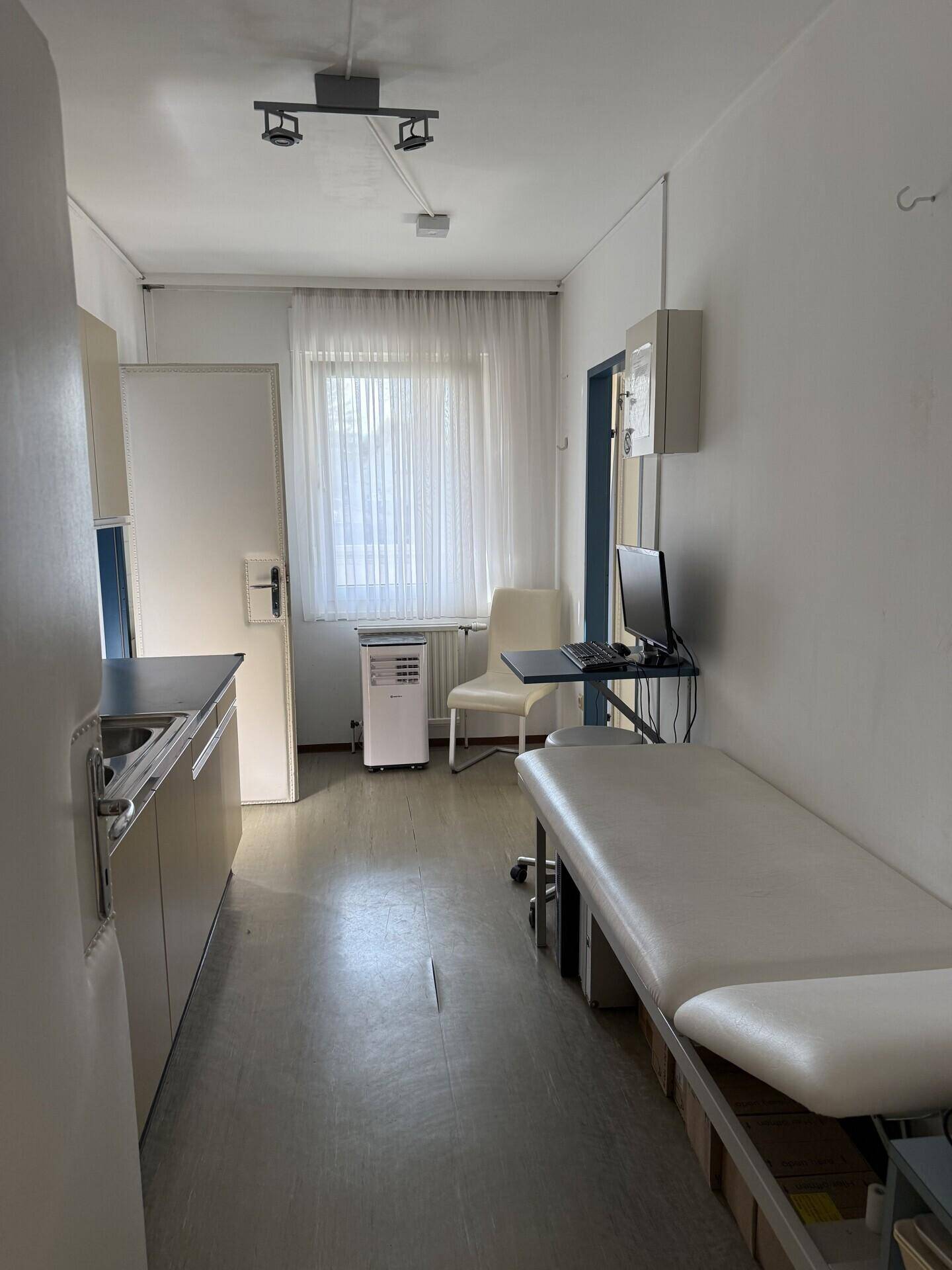 Kleines Behandlungszimmer mit Untersuchungsliege, Schreibtisch und Fenster mit Vorhängen.