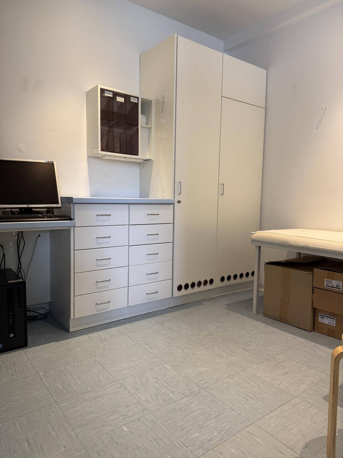 Büroraum mit weißen Schränken, Schubladen und einer Untersuchungsliege an der Wand.