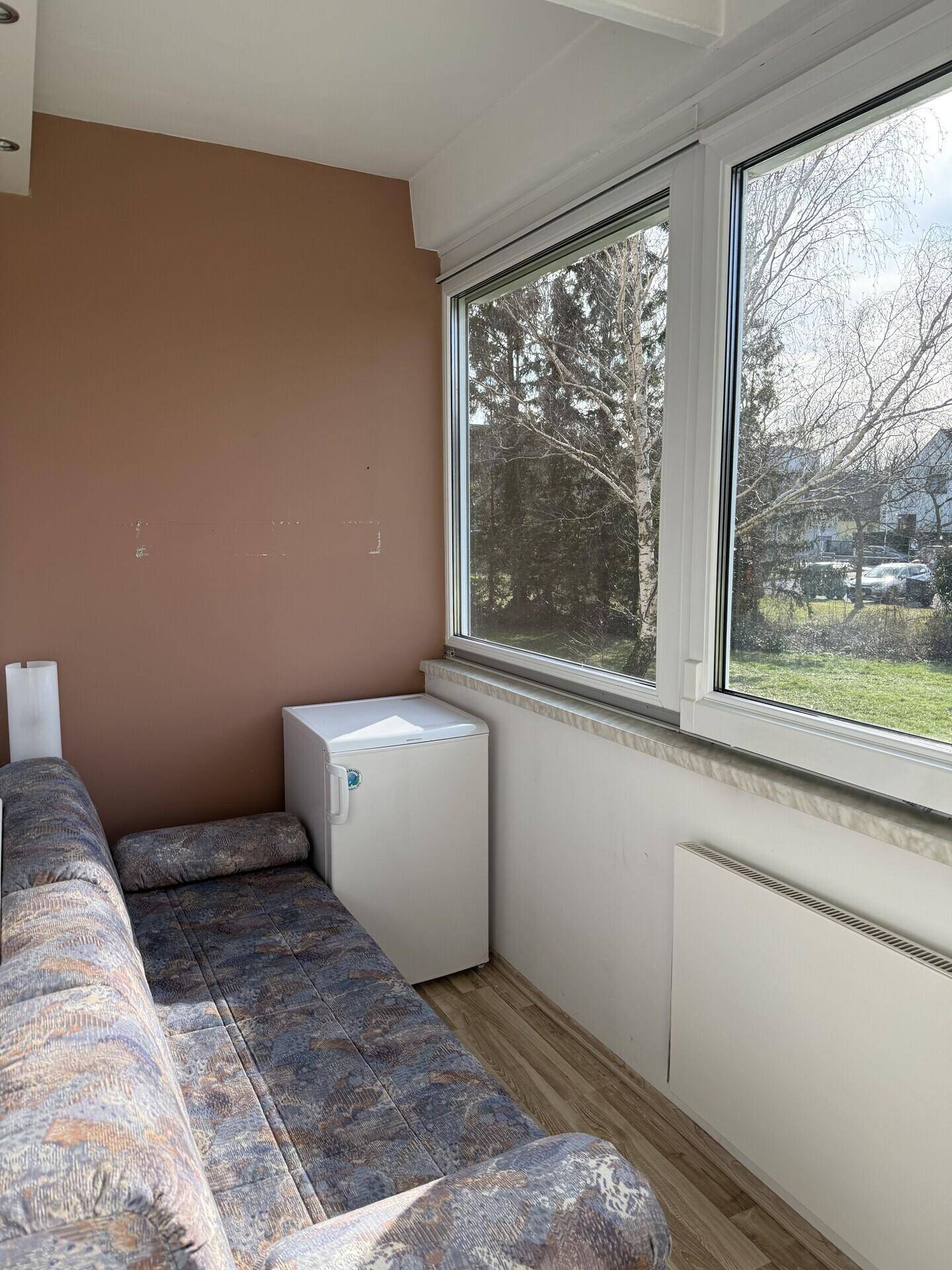 Verglaster Balkon oder Wintergarten mit Sofa, kleinem Kühlschrank und Blick ins Grüne.