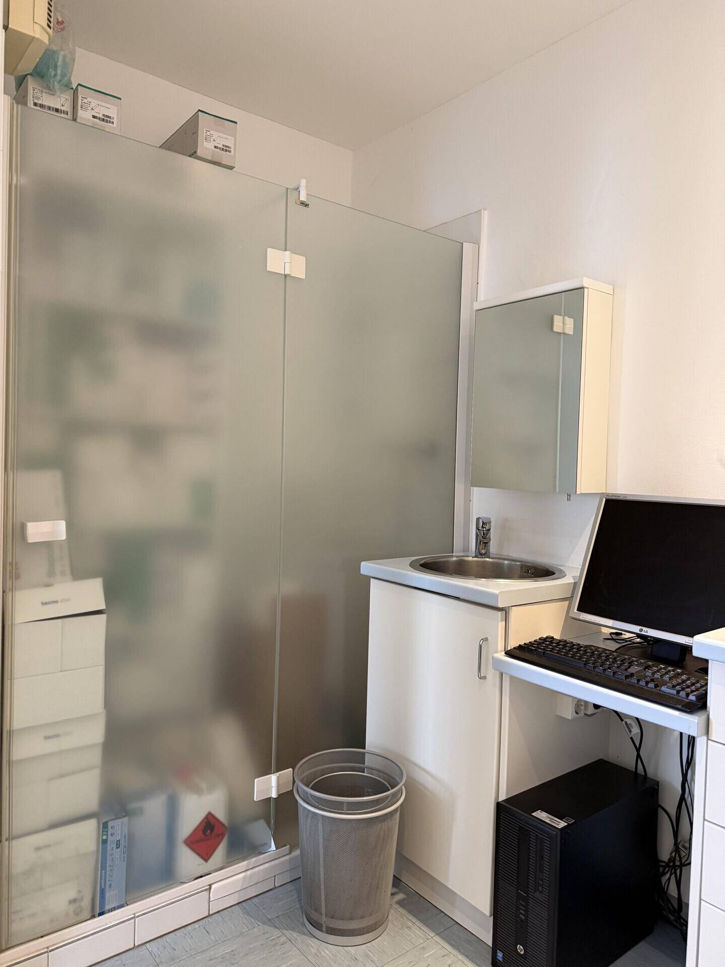 Kleiner Raum mit Waschbecken, Computer und einer Milchglasabtrennung für Privatsphäre.