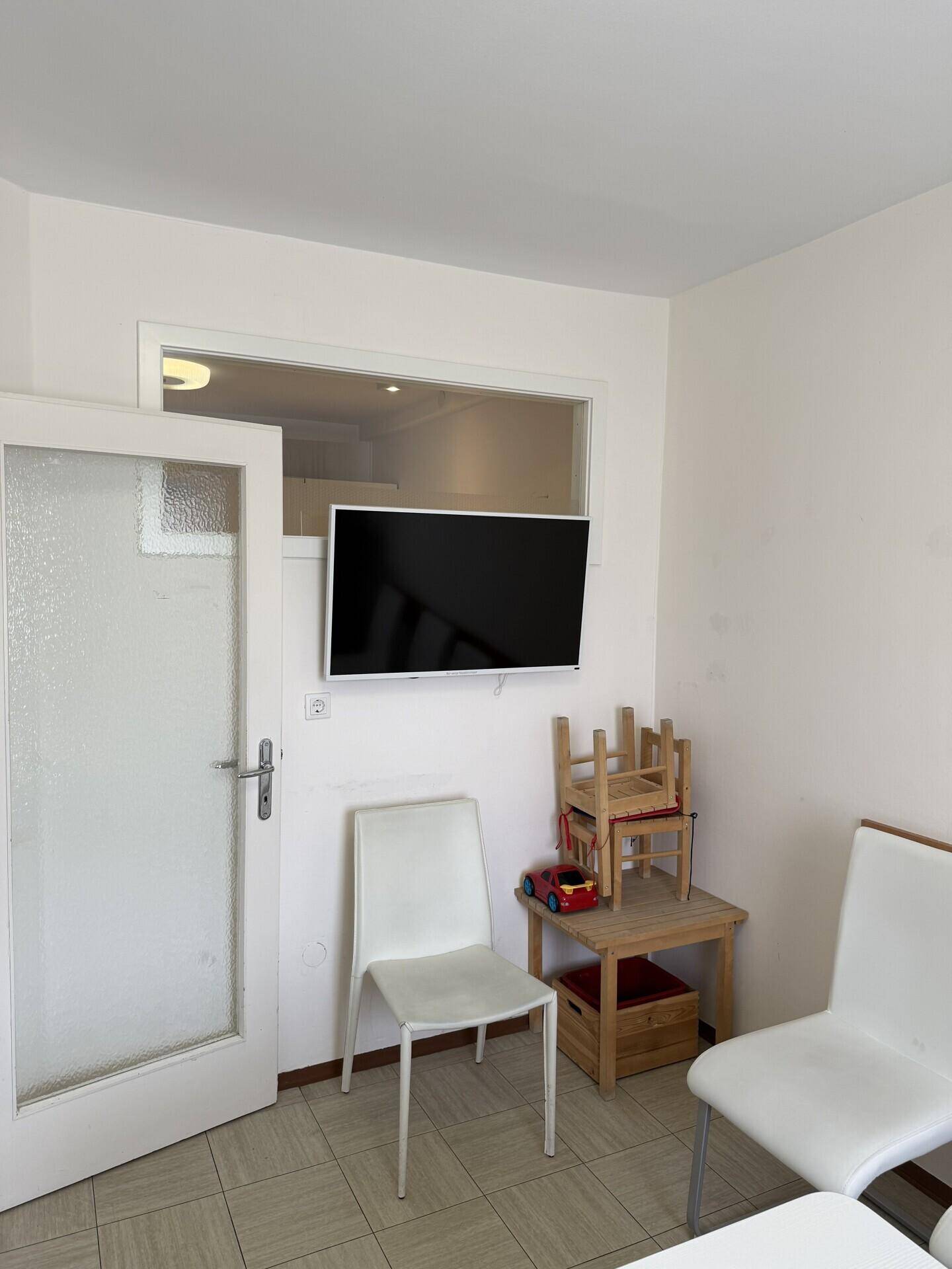 Wartebereich oder kleines Büro mit Stühlen, Holztisch und einem an der Wand montierten Fernseher.