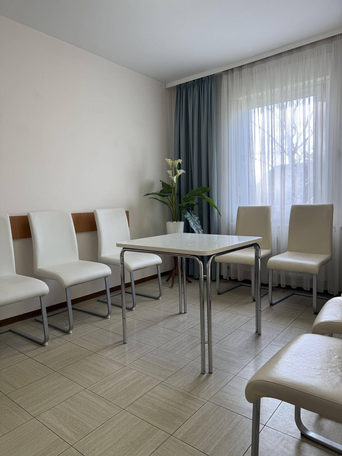 Wartebereich mit mehreren Stühlen, einem Tisch und einem großen Fenster mit Vorhängen.