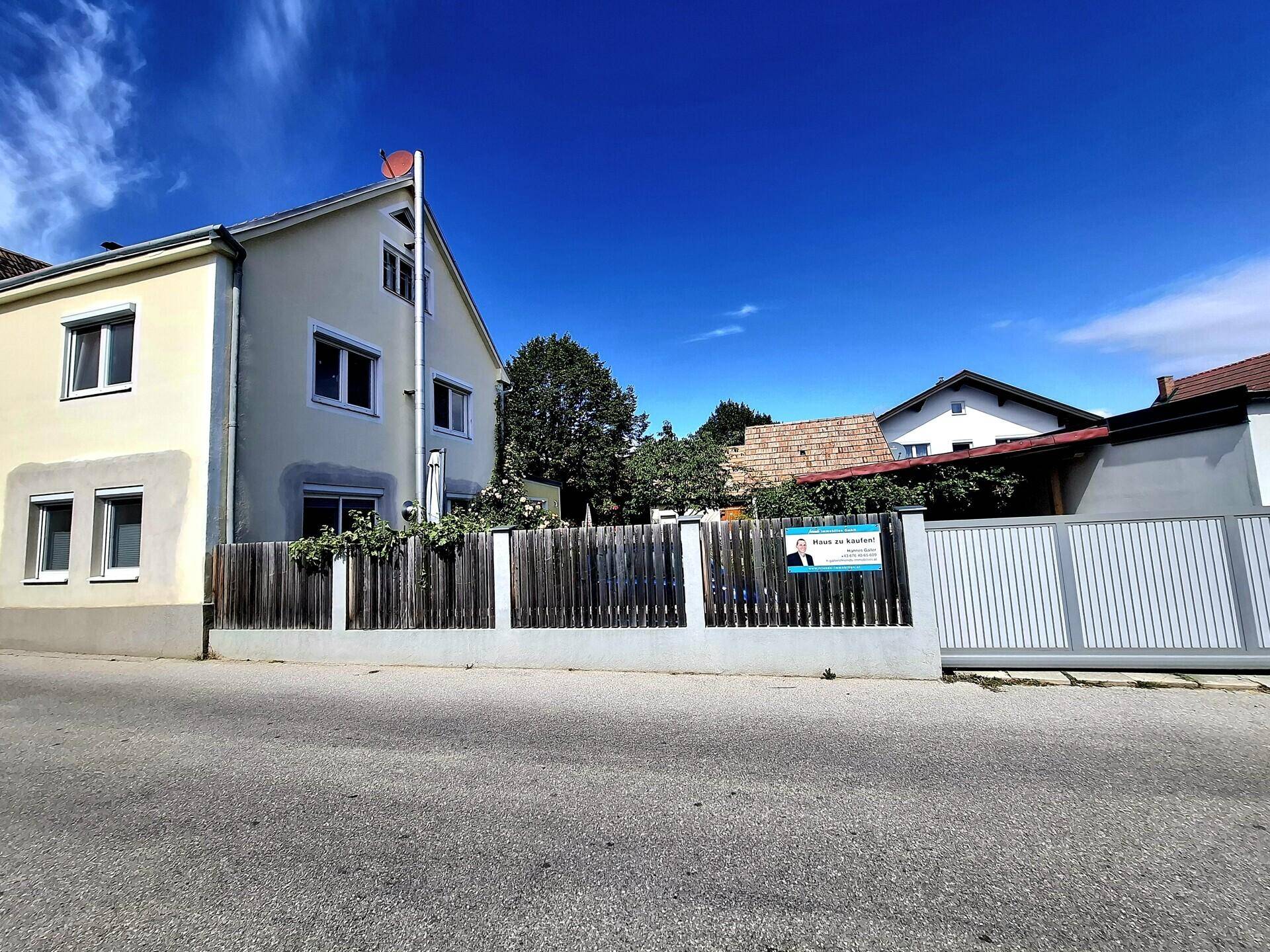 Straßenansicht eines Hauses mit Holzzaun und einem Verkaufsschild an einem sonnigen Tag.