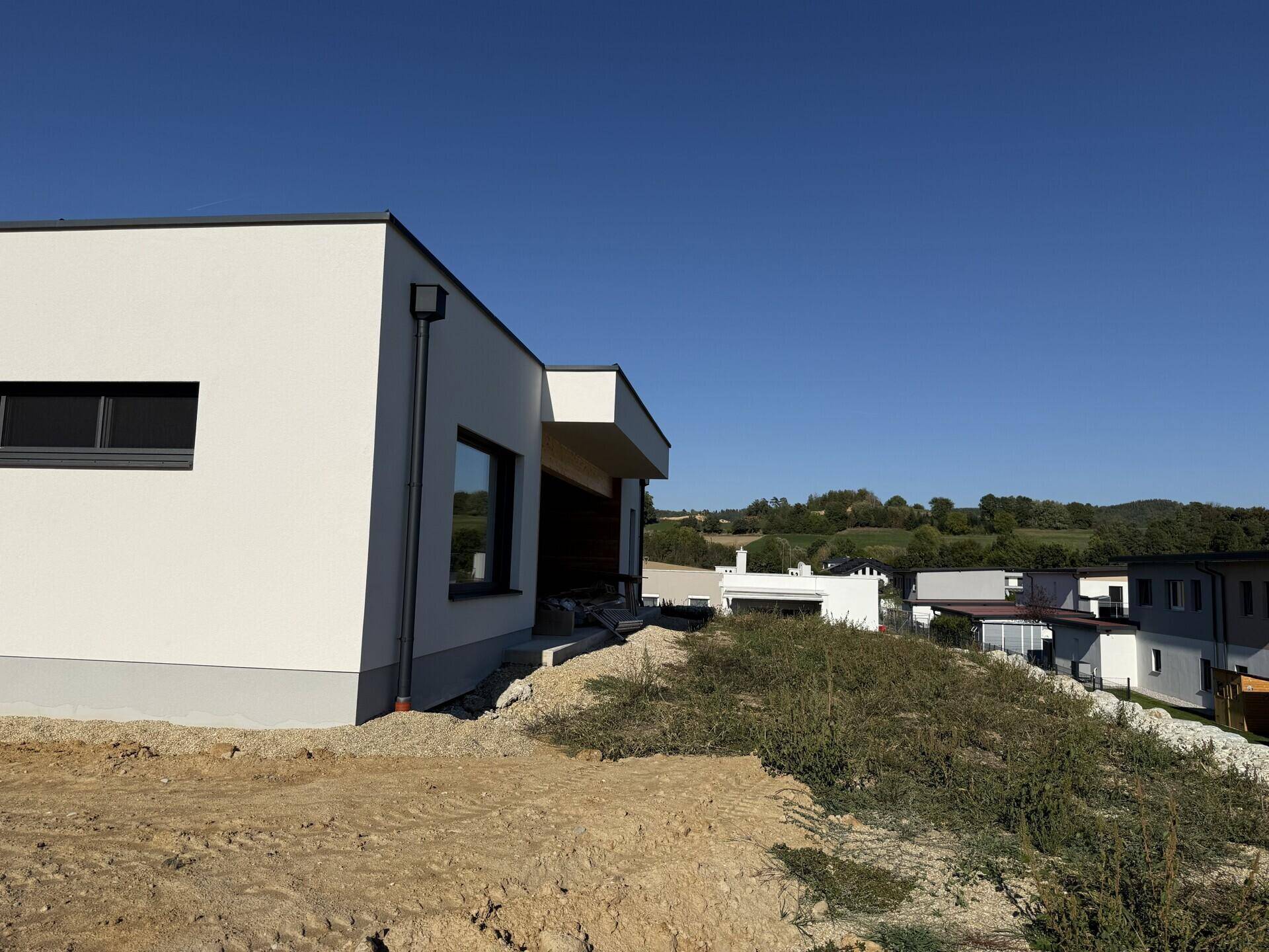 Seitenansicht eines modernen Neubaus mit weißer Fassade und angrenzendem unbebautem Grundstück.