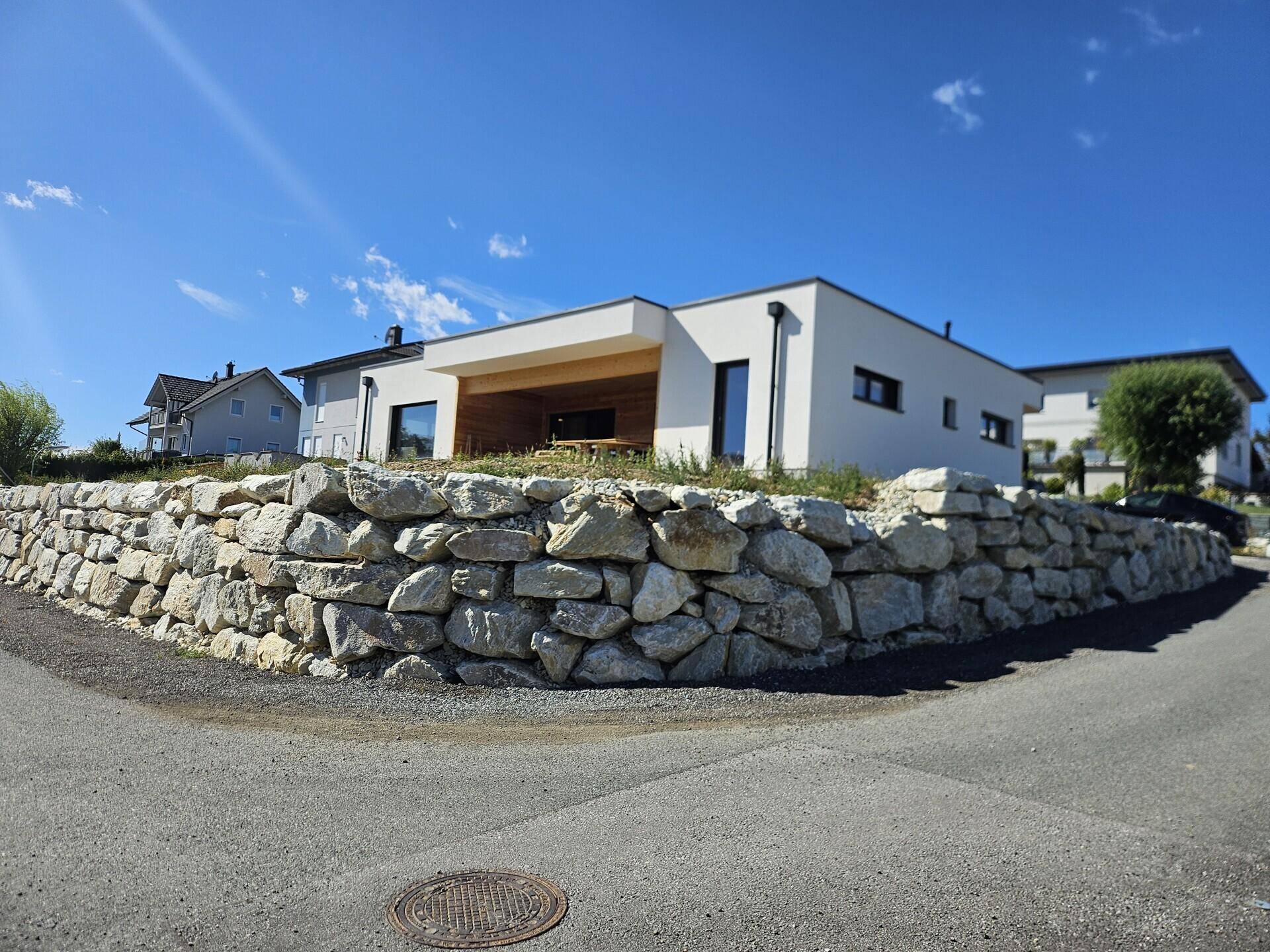 Modernes Einfamilienhaus mit weißer Fassade und einer Natursteinmauer im Vordergrund unter blauem Himmel.