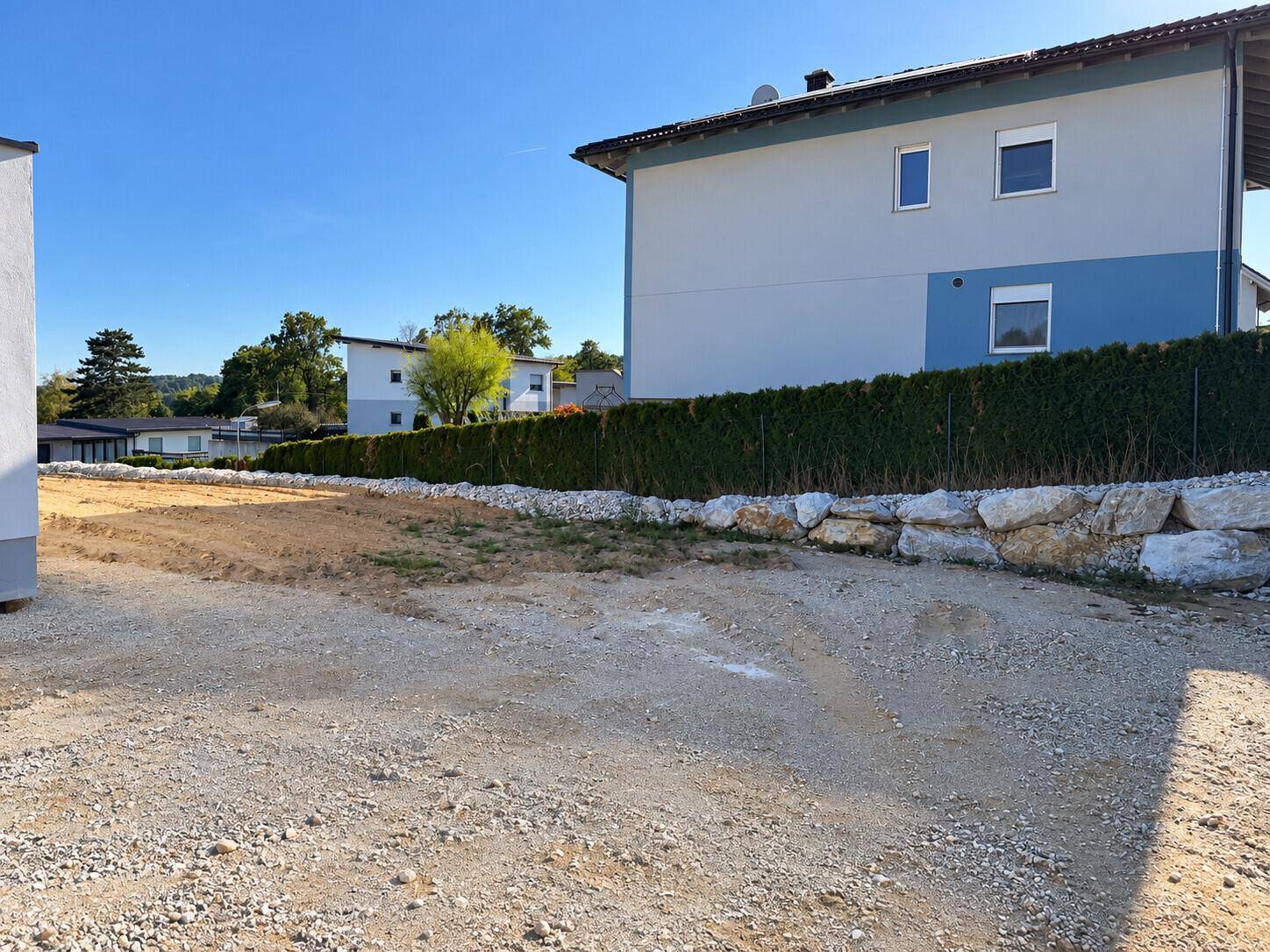 Unbebautes Grundstück mit Hecke und angrenzenden Wohnhäusern in ländlicher Umgebung.