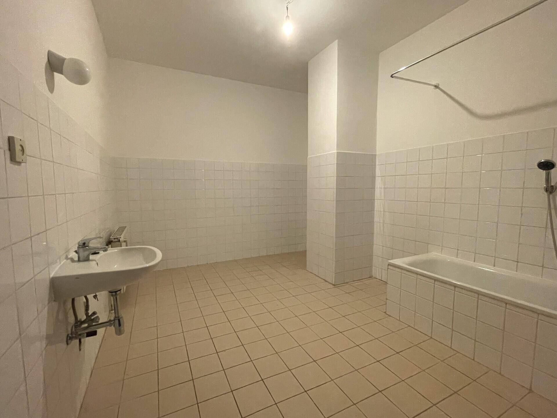 Badezimmer mit weißen Wandfliesen, beigefarbenem Fliesenboden, Waschbecken und Badewanne mit Duschfunktion.