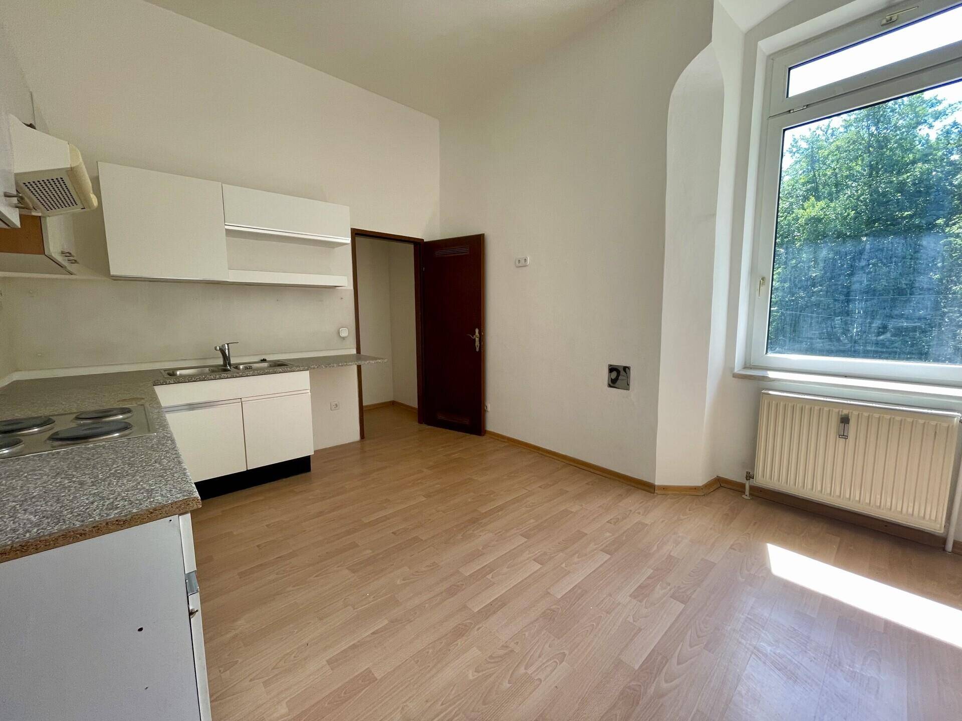 Küche mit weißen Einbauschränken, Herd und Spüle, dazu Laminatboden und Fensterblick ins Grüne.