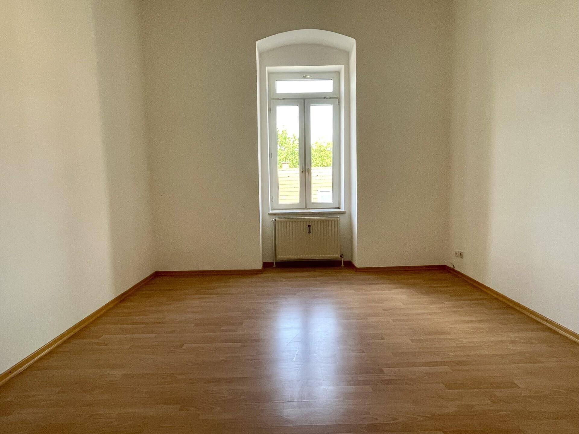 Leeres Zimmer mit hellem Laminatboden, weißen Wänden und einem großen Fenster mit Heizkörper.