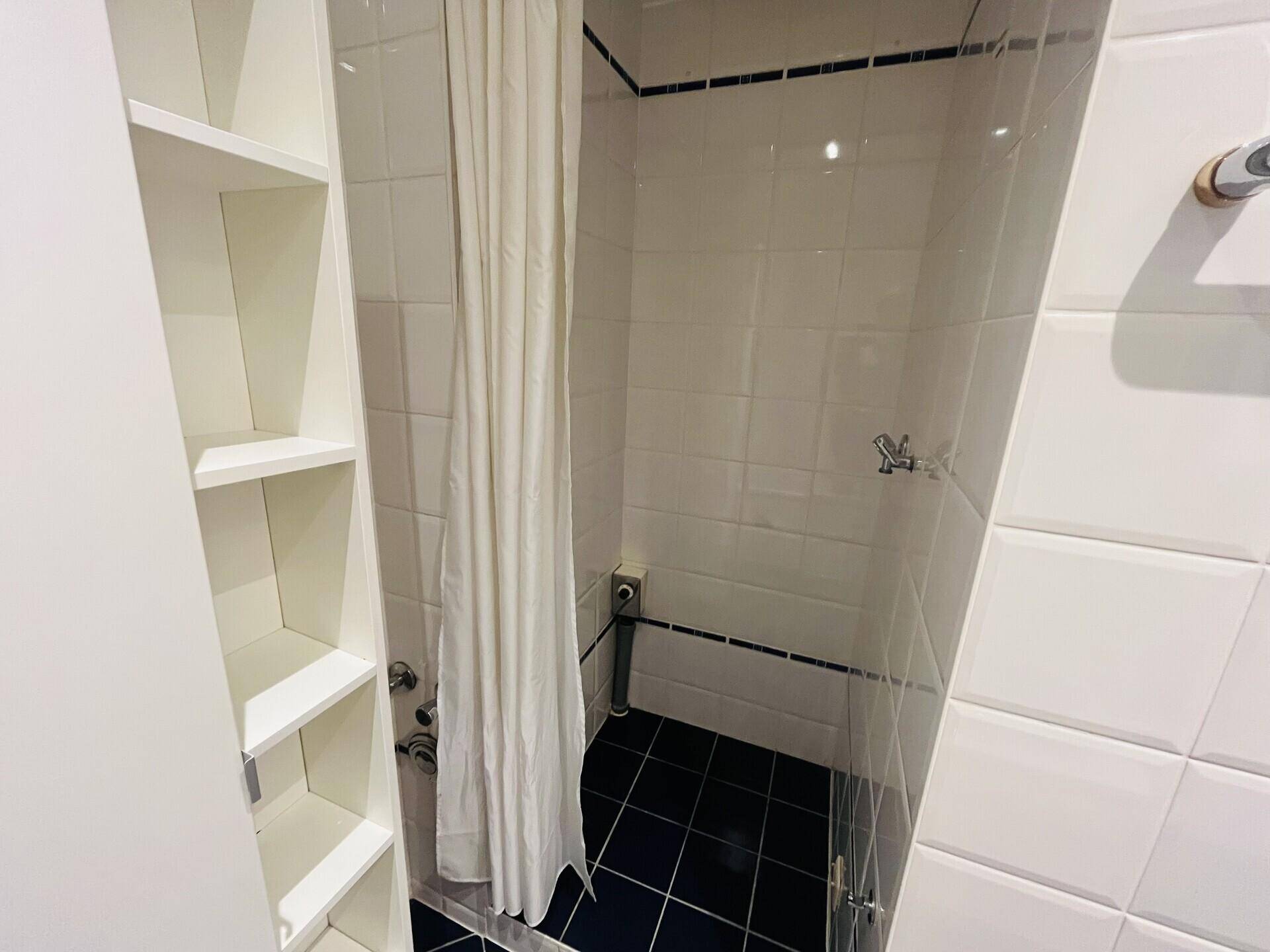 Duschbereich im Badezimmer mit weißen Fliesen, dunklem Boden und Duschvorhang.
