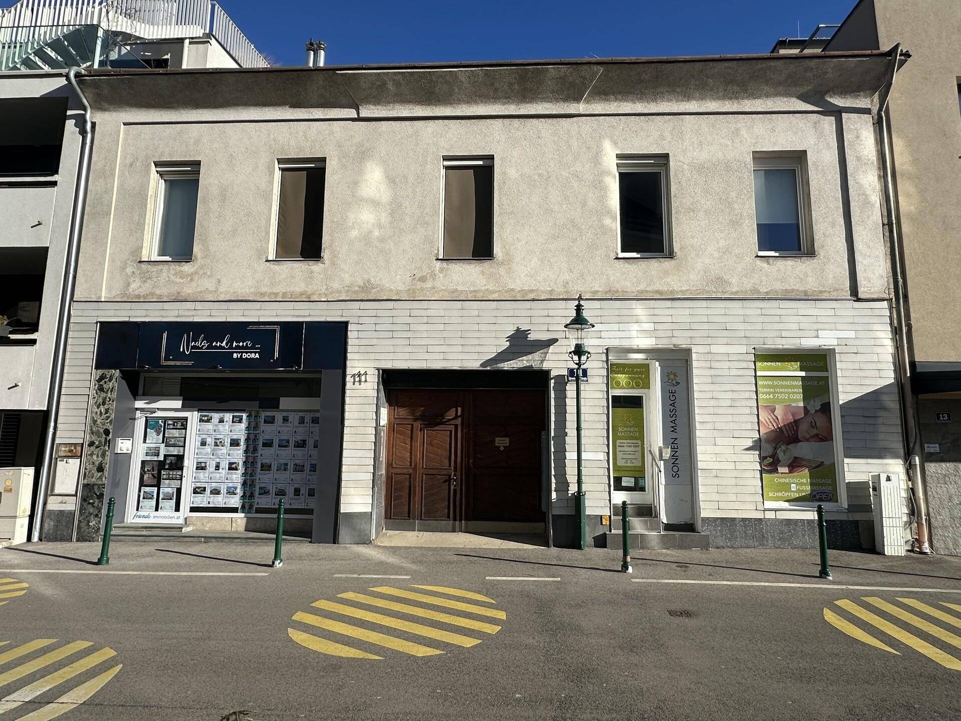 Fassade eines älteren Gebäudes mit Geschäften im Erdgeschoss und Wohnungen darüber an einer Straße.