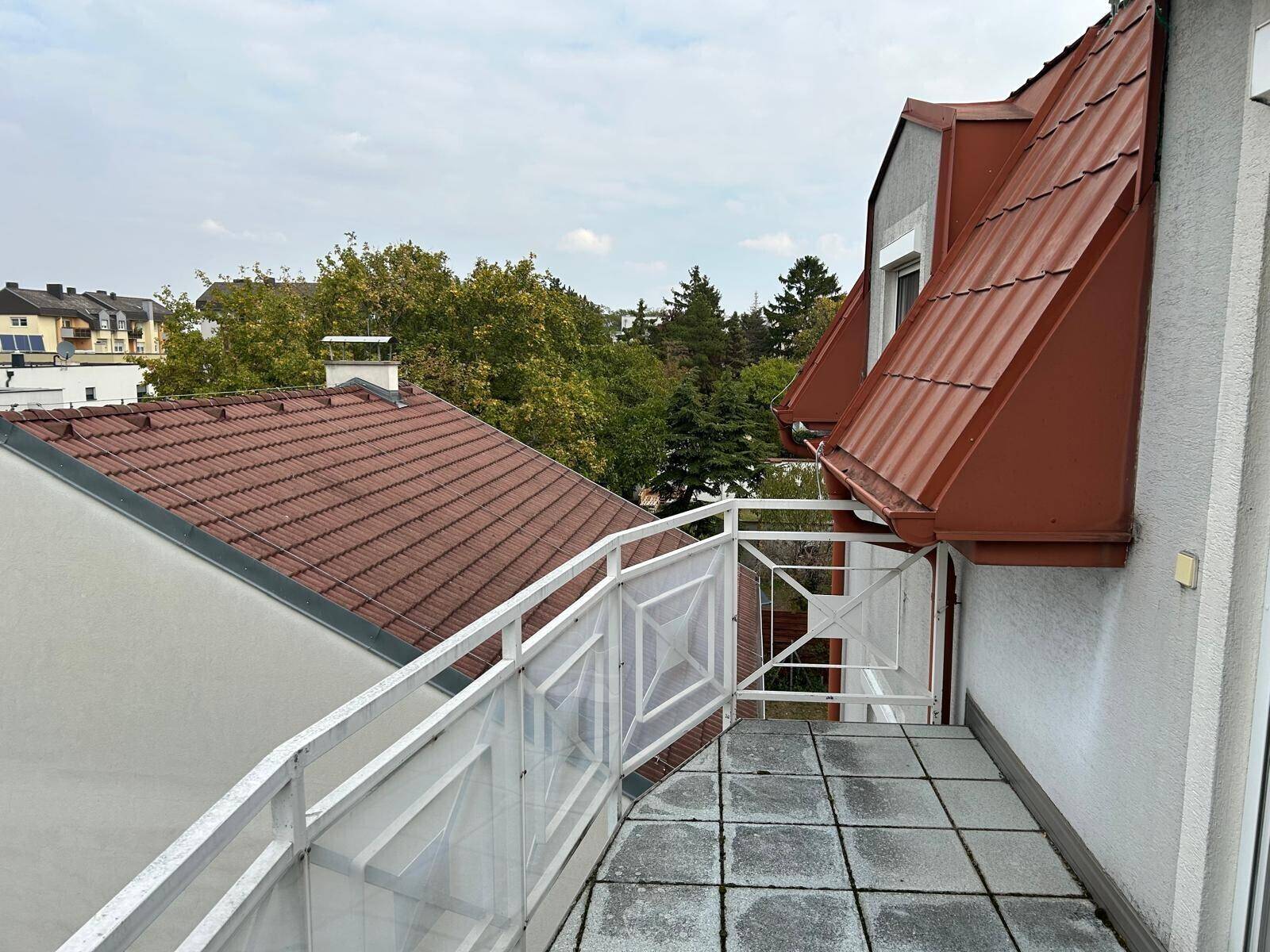 Balkon mit Fliesenboden und Blick auf die umliegenden Dächer und Bäume.