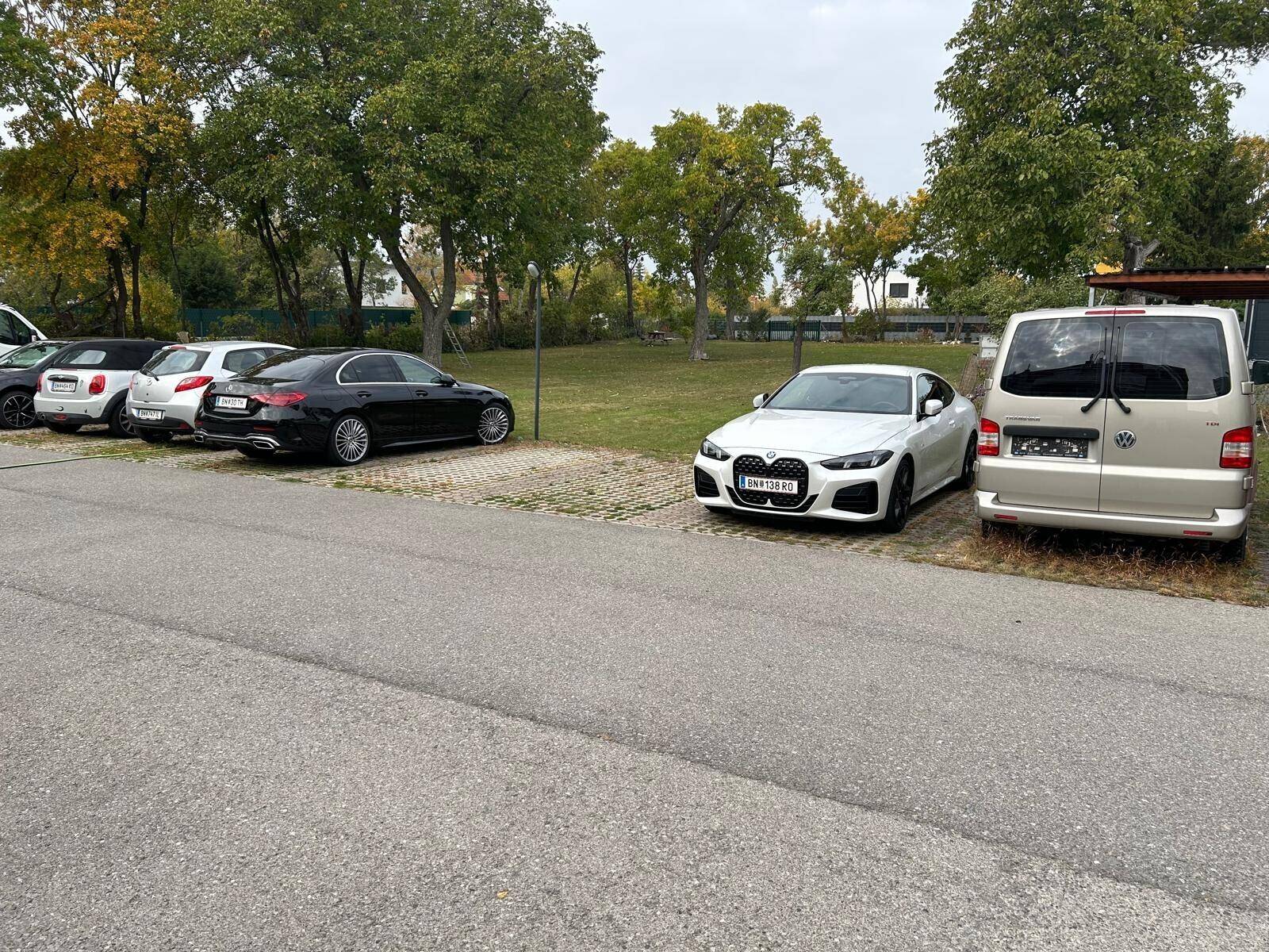 Außenparkplatz mit mehreren Autos und Bäumen im Hintergrund unter bewölktem Himmel.