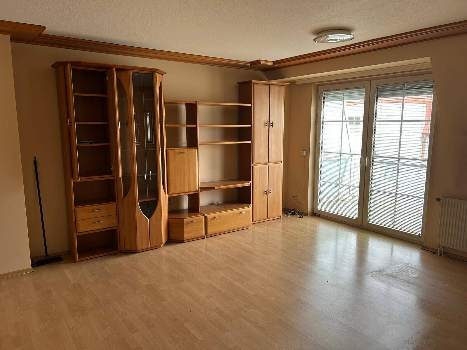 Geräumiges Wohnzimmer mit Holzschrankwand und Zugang zu einem Balkon durch große Fenstertüren.