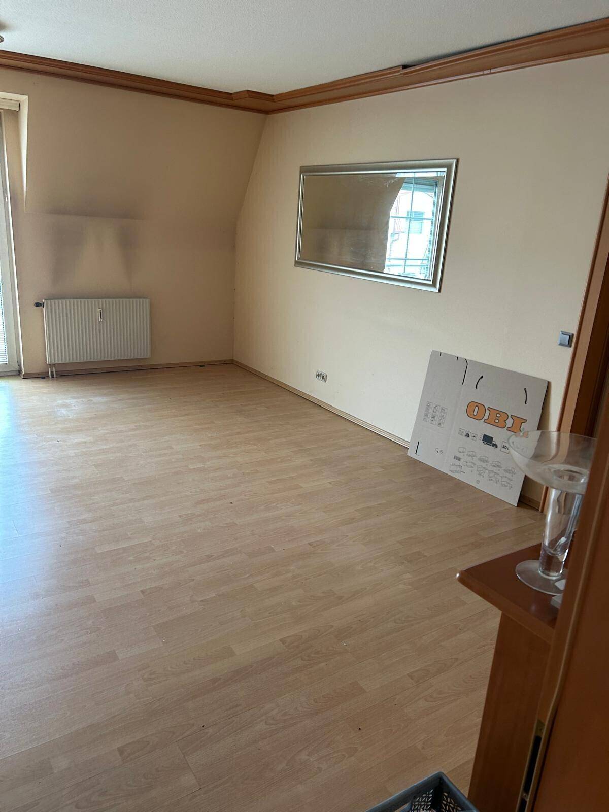 Leeres Wohnzimmer mit Holzboden, schrägen Wänden und einem großen Spiegel an der Wand.