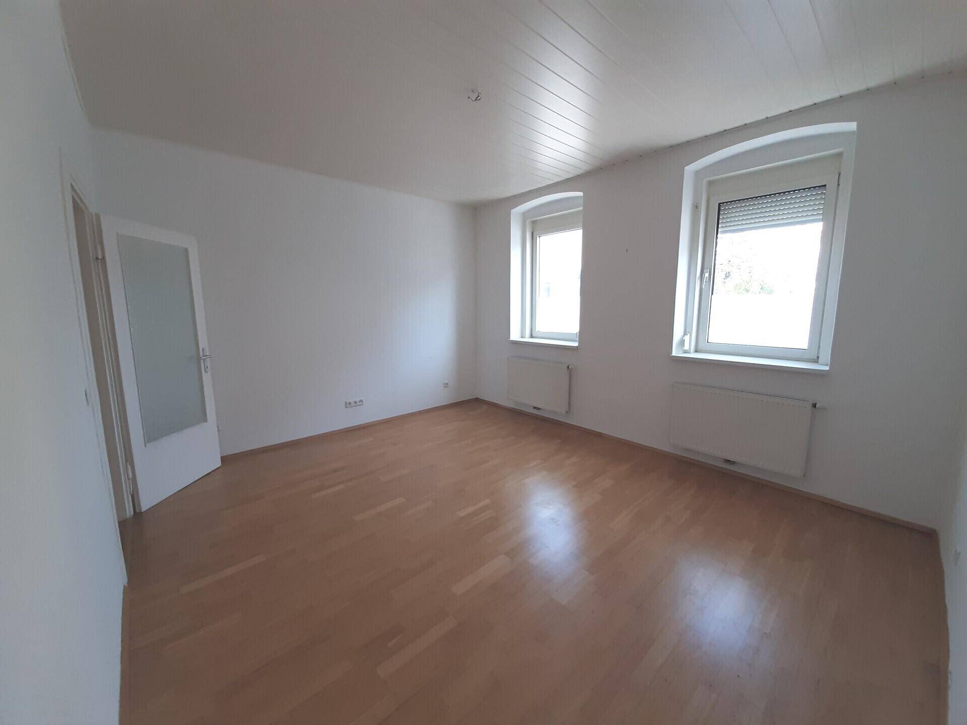 Helles Zimmer mit Parkettboden, zwei Fenstern und weiß gestrichenen Wänden.
