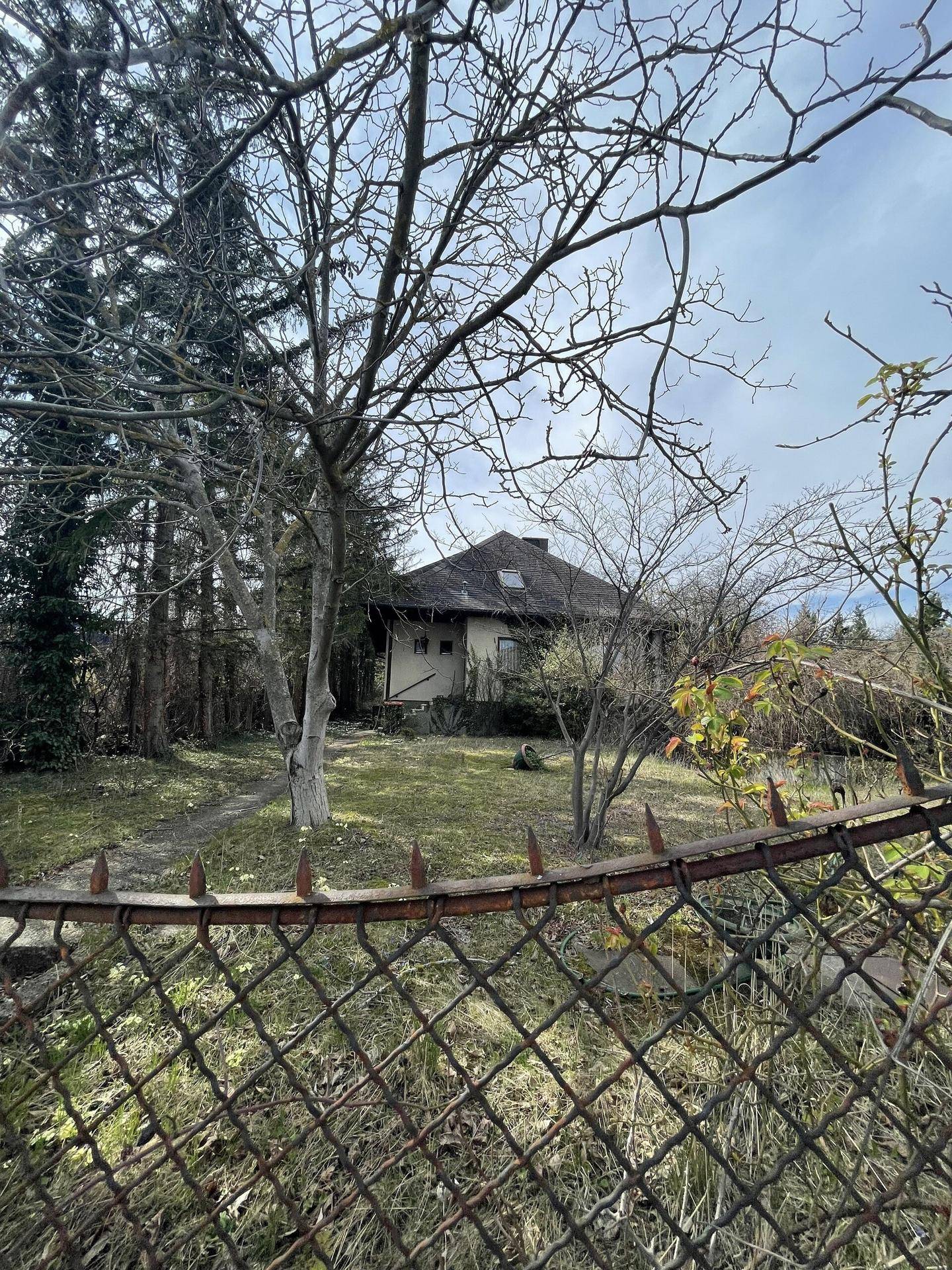 Blick auf das Haus durch einen rostigen Zaun, umgeben von kahlen Bäumen und einem verwilderten Garten.