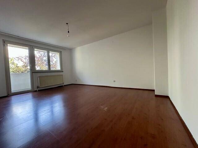 Helles, leeres Zimmer mit Laminatboden, weißen Wänden und Zugang zu einem Balkon.