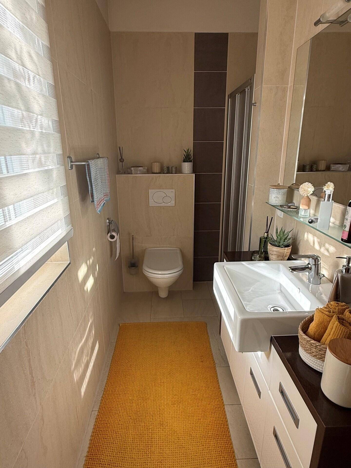 Modernes Badezimmer mit beigen Fliesen, wandhängendem WC, Dusche und Fenster für Tageslicht.