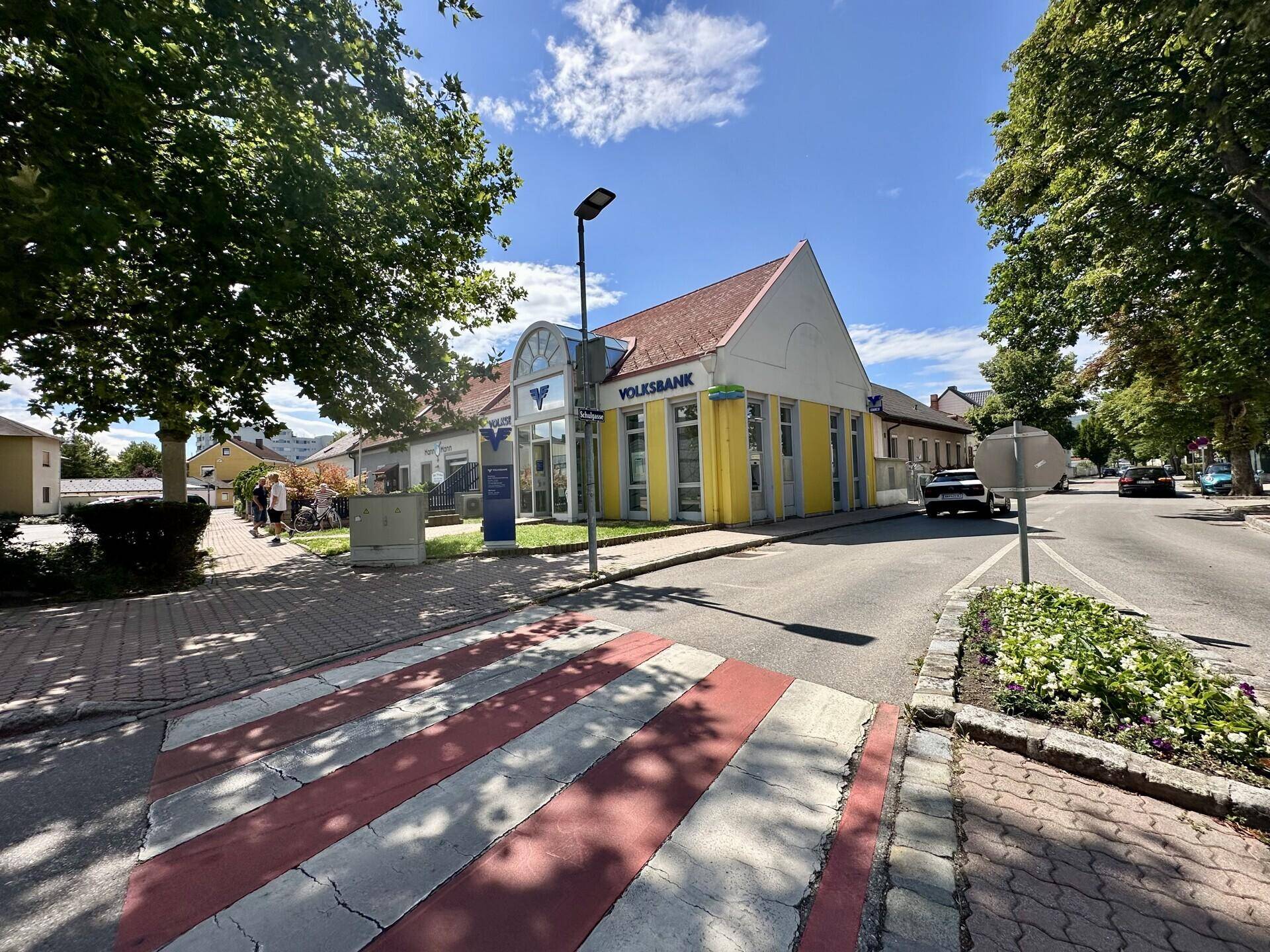 Außenansicht eines Bankgebäudes an einer belebten Straße mit Zebrastreifen und Bäumen.