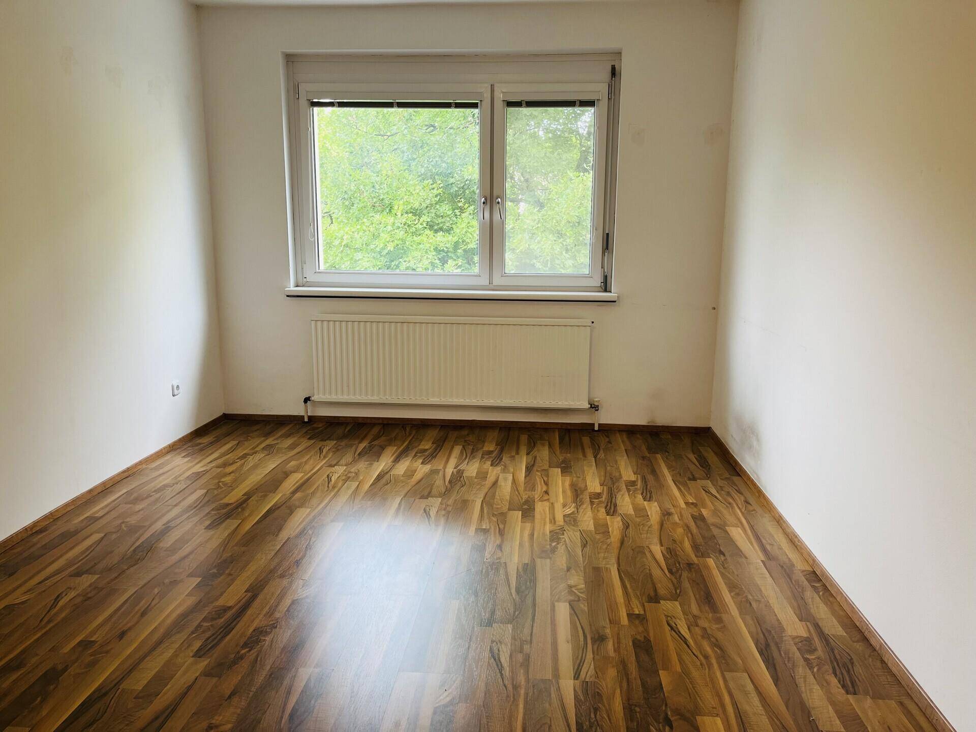 Schlafzimmer mit Laminatboden, hellen Wänden und Fenster mit Grünblick.