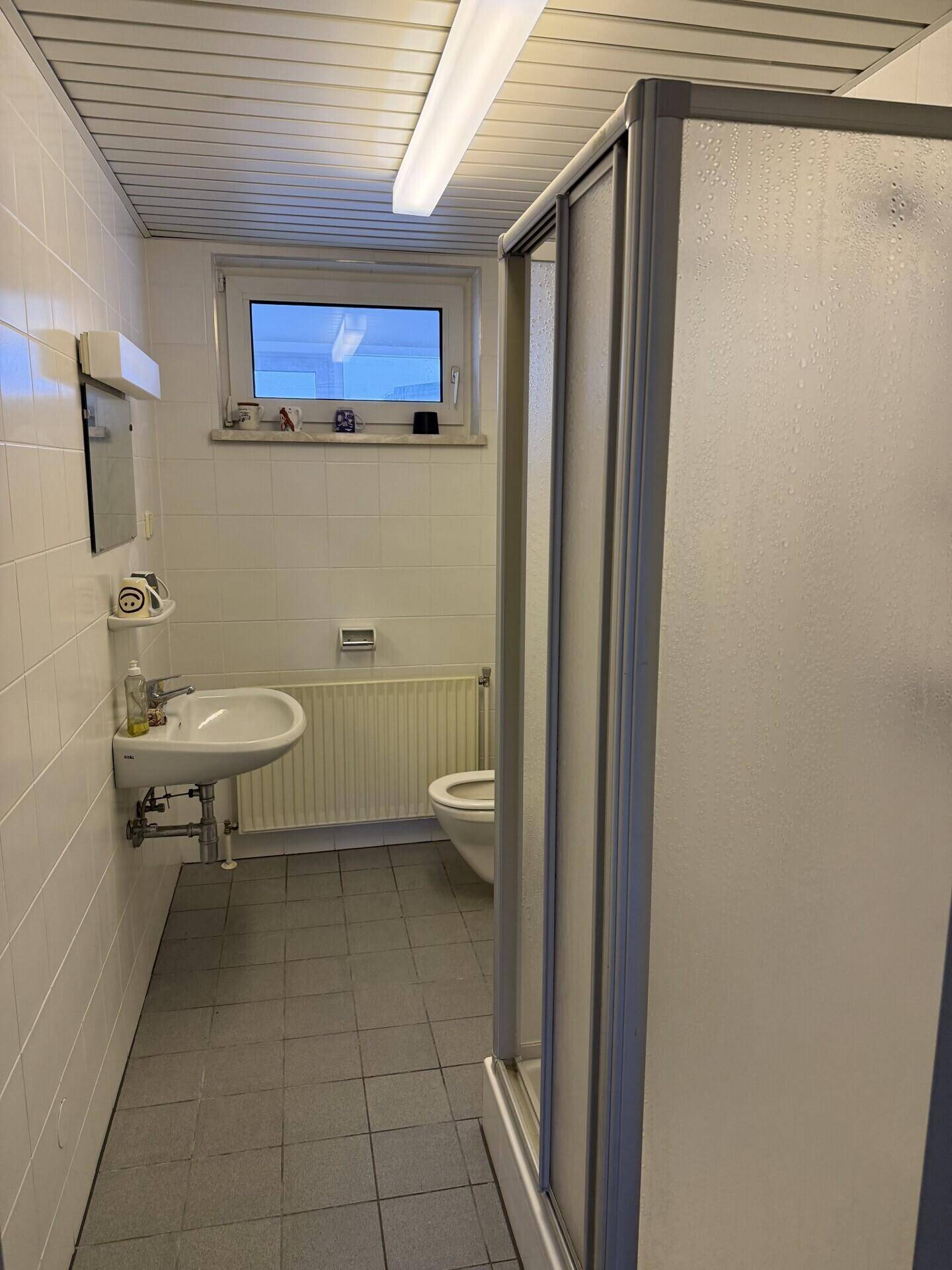 Badezimmer mit weißen Fliesen, einer Duschkabine, Toilette, Waschbecken und einem Fenster.